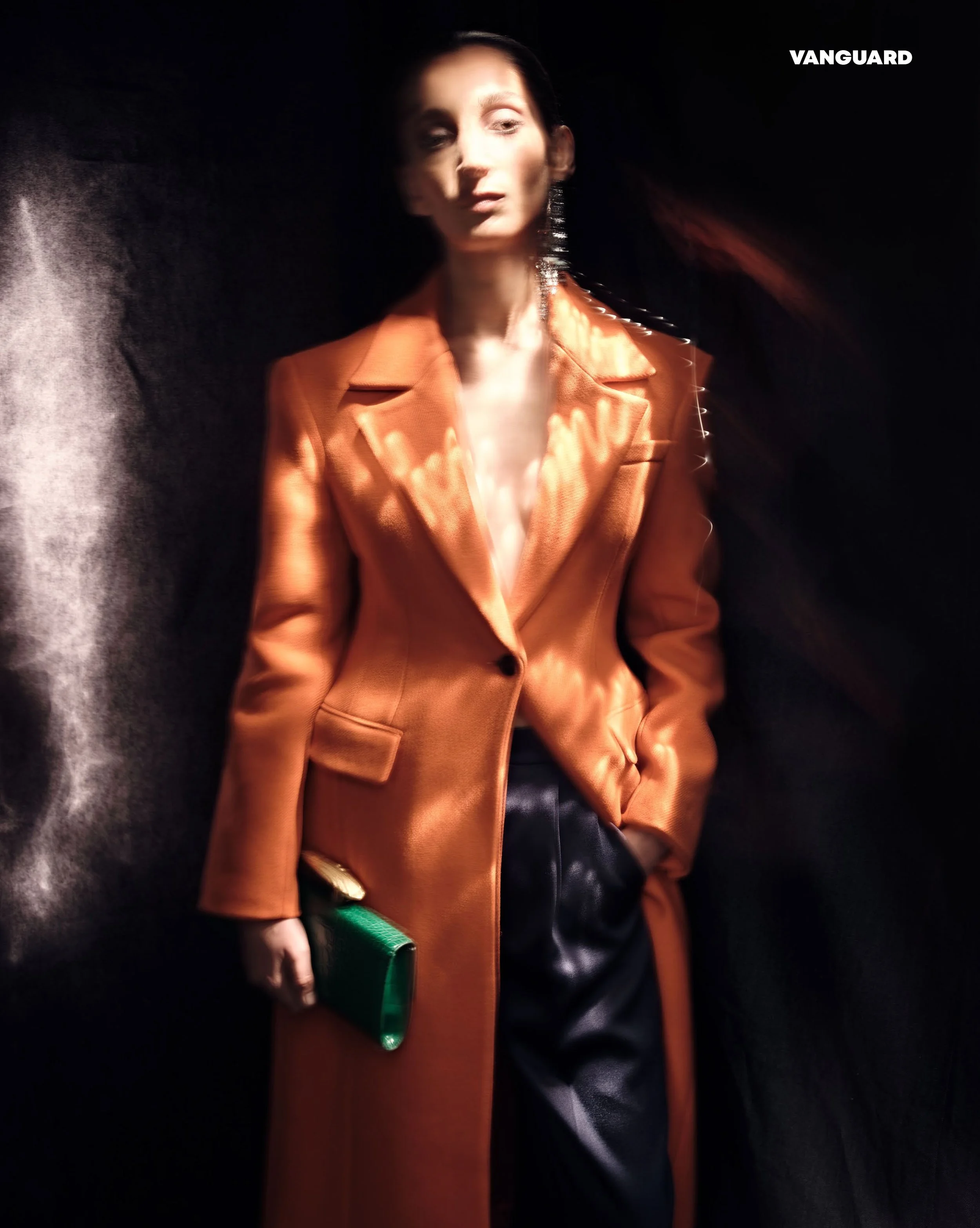 A woman in an orange blazer with a black collar, holding a green clutch, standing against a black background with the word 'VANGUARD' in white text in the top right corner.