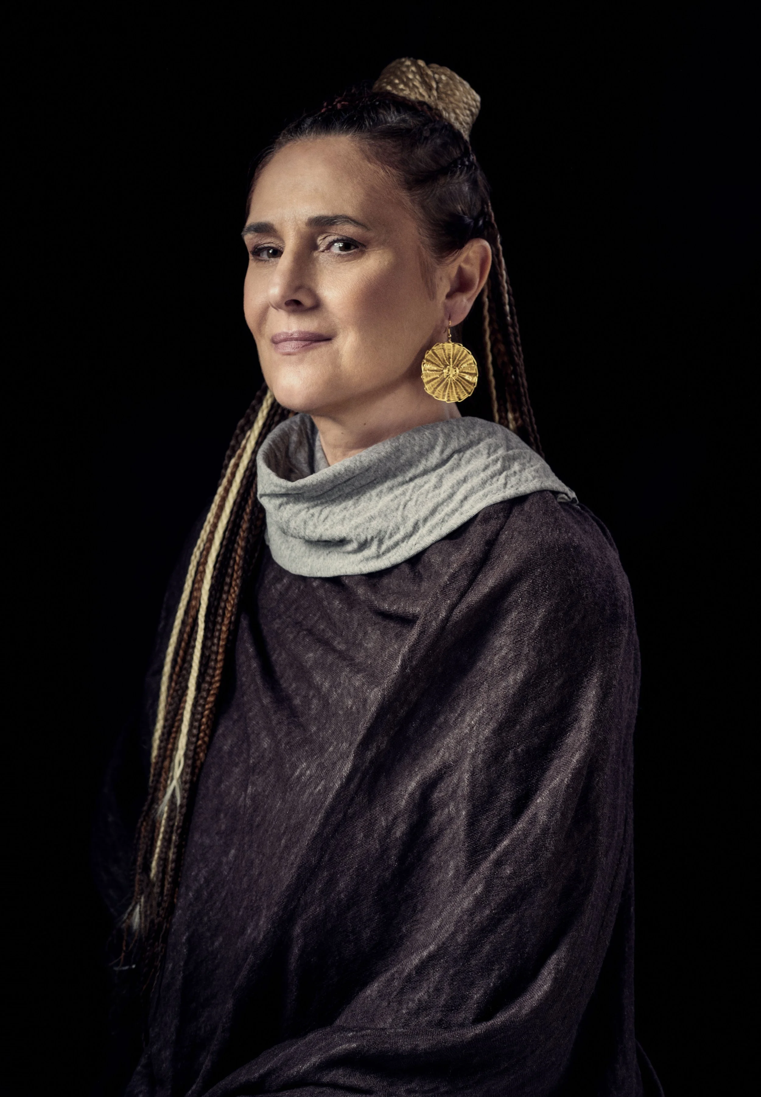 A woman with long braids and a beige bun hairstyle, wearing large round gold earrings, a gray scarf around her neck, and a dark jacket, against a black background.