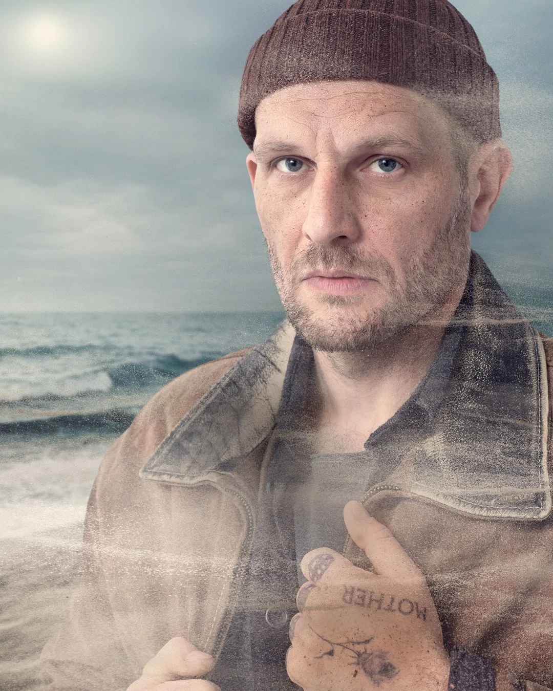 A man with light skin, blue eyes, a beard, and freckles wears a brown beanie and a brown leather jacket with a zipper. He is standing outdoors with a cloudy sky and ocean behind him, holding the collar of his jacket with one hand.