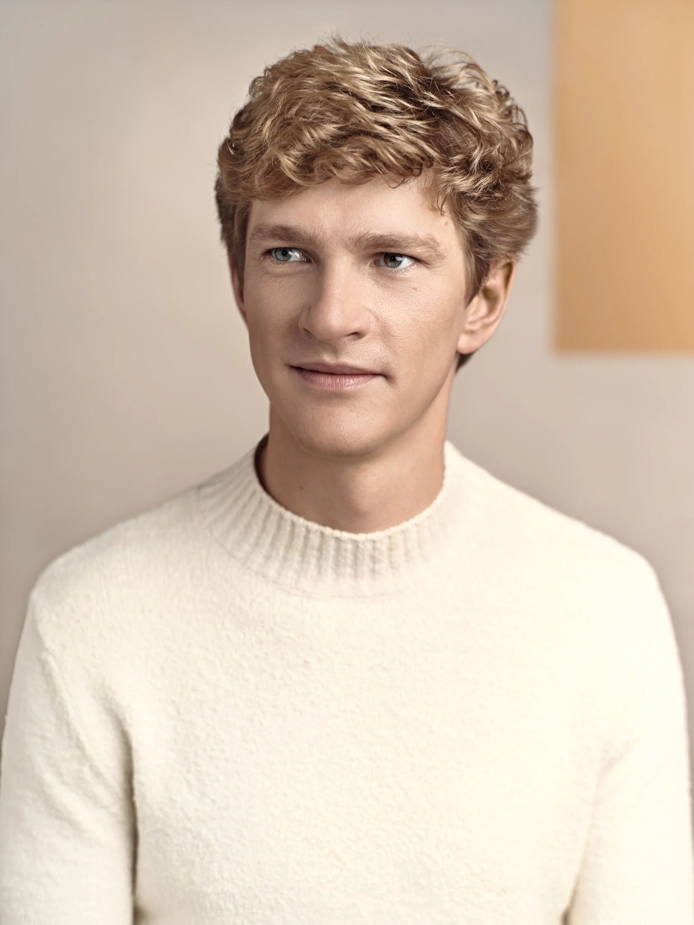 Close-up of a young man's face with blonde, slightly wavy hair, wearing a white knit sweater, against a neutral background.
