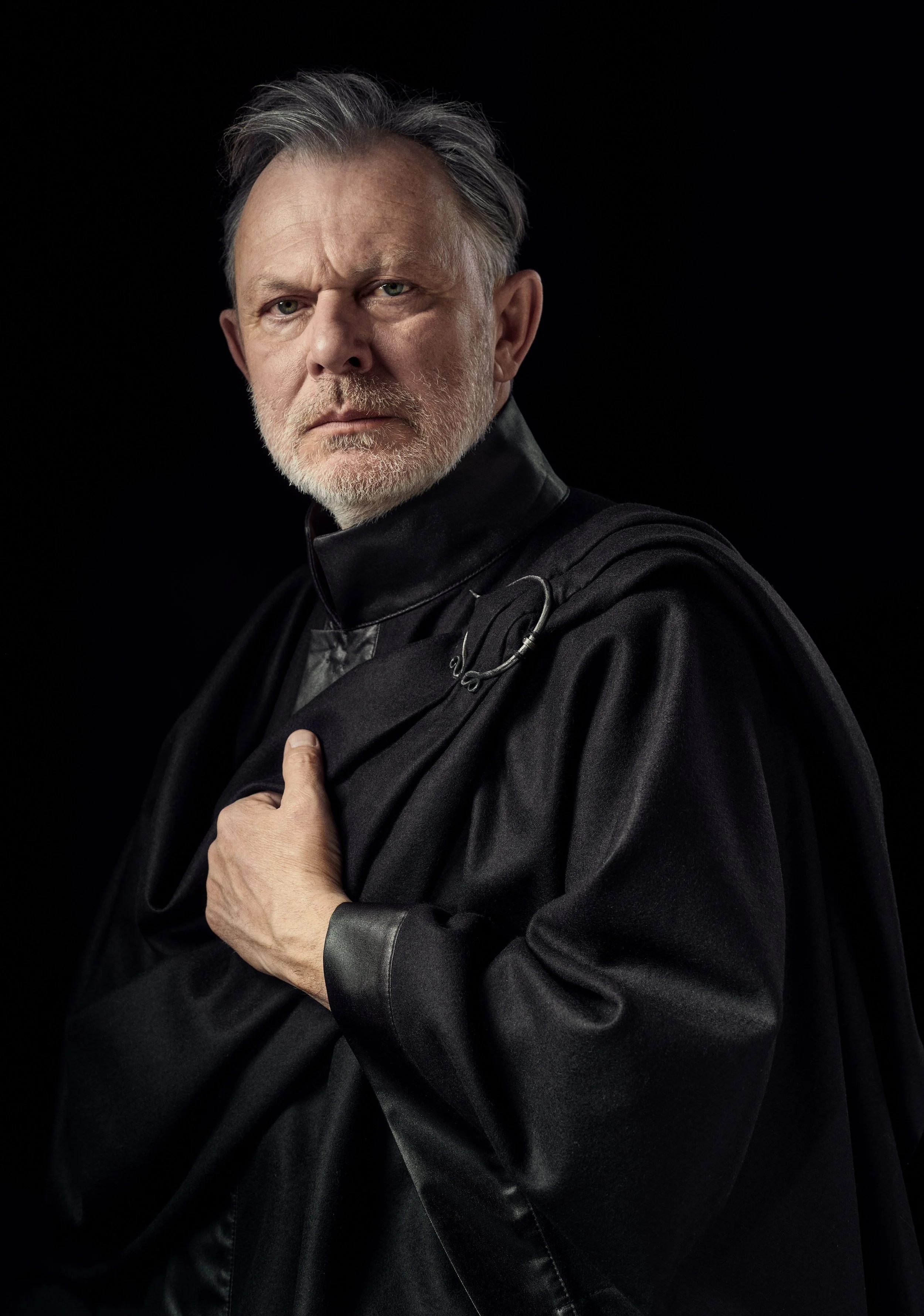 A middle-aged man with gray hair and beard wearing a black leather jacket with a high collar, looking serious, against a black background.