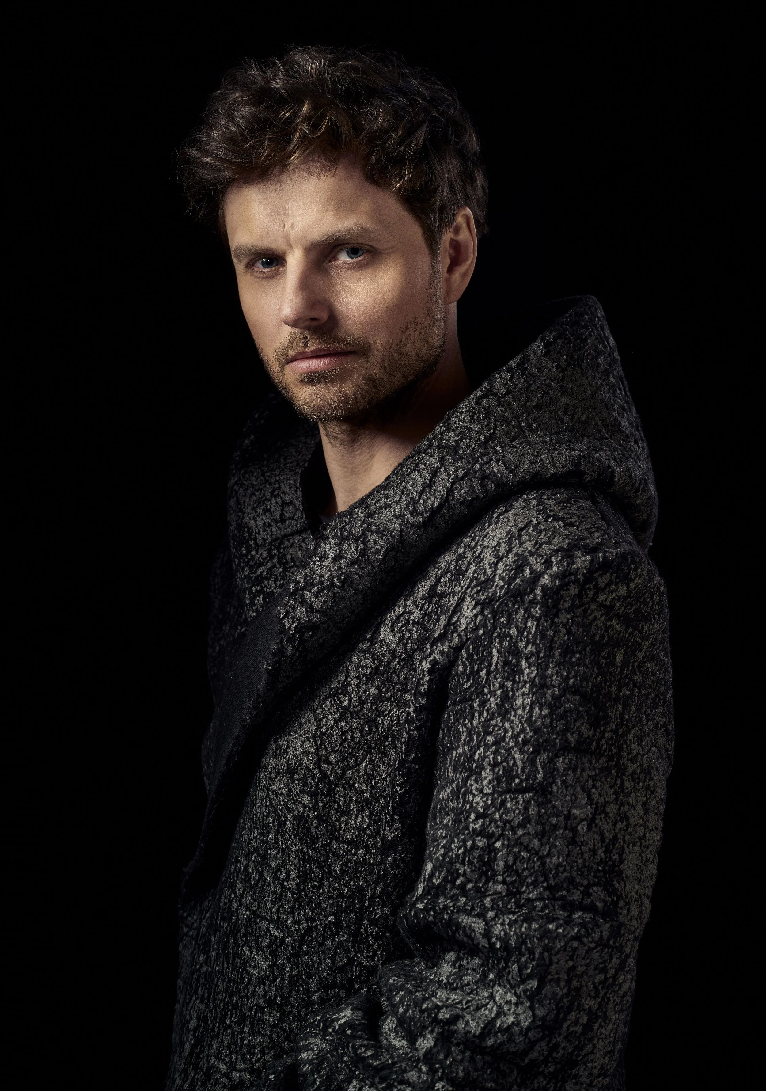 A man with light skin, brown curly hair, and a beard, wearing a textured dark gray hoodie, looking into the camera, against a black background.