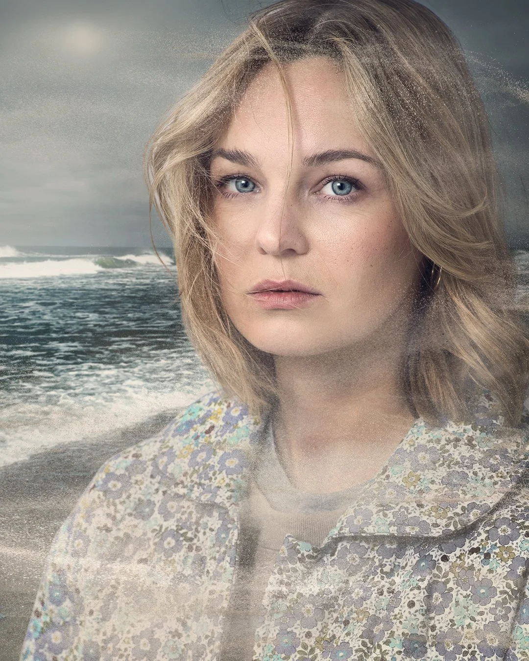 A woman with blue eyes and shoulder-length blonde hair standing in front of a stormy ocean scene, with waves and cloudy sky, wearing a floral patterned shirt.
