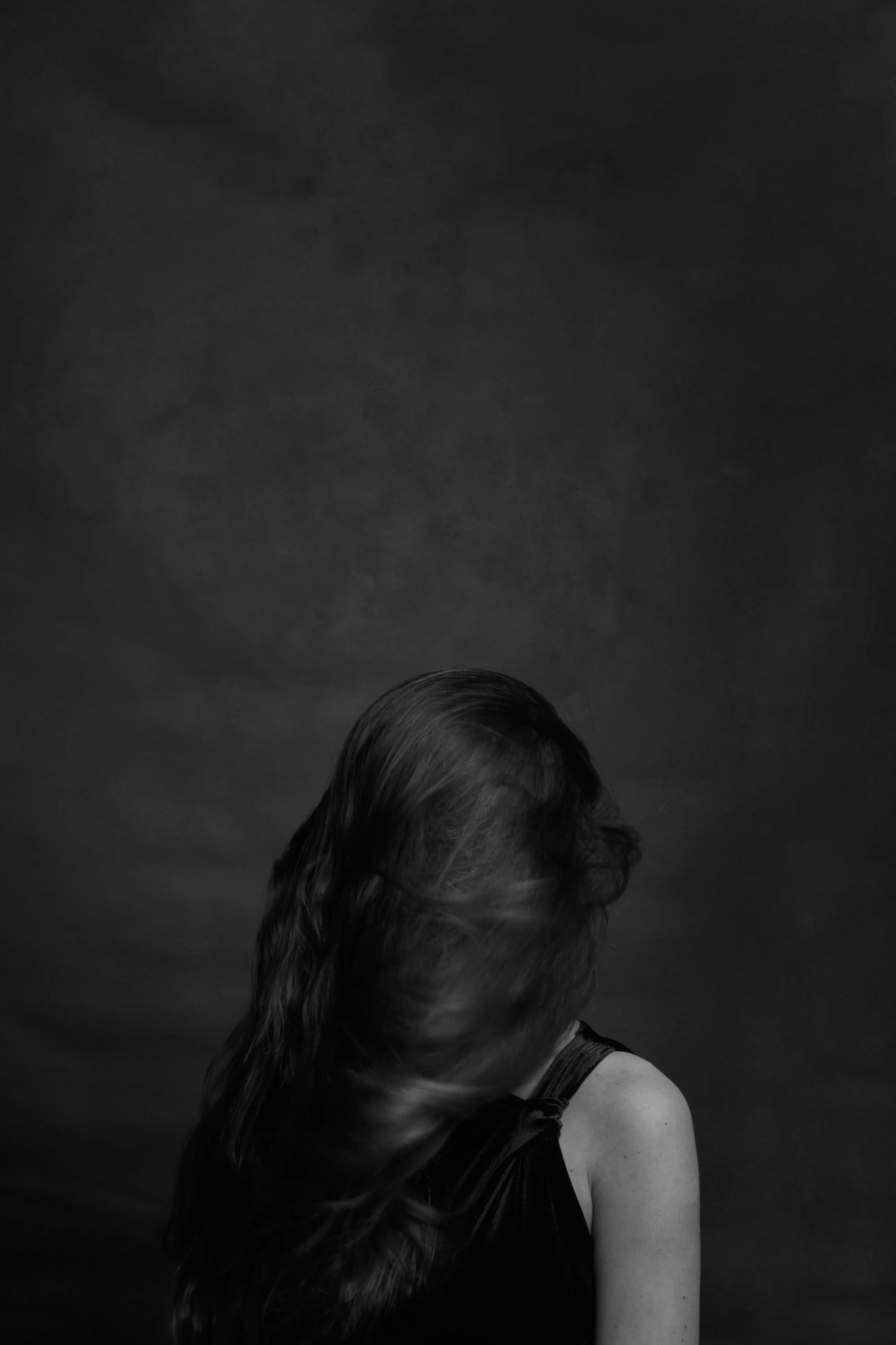 A black and white photograph of a woman with long wavy hair, partially covering her face, against a dark background.