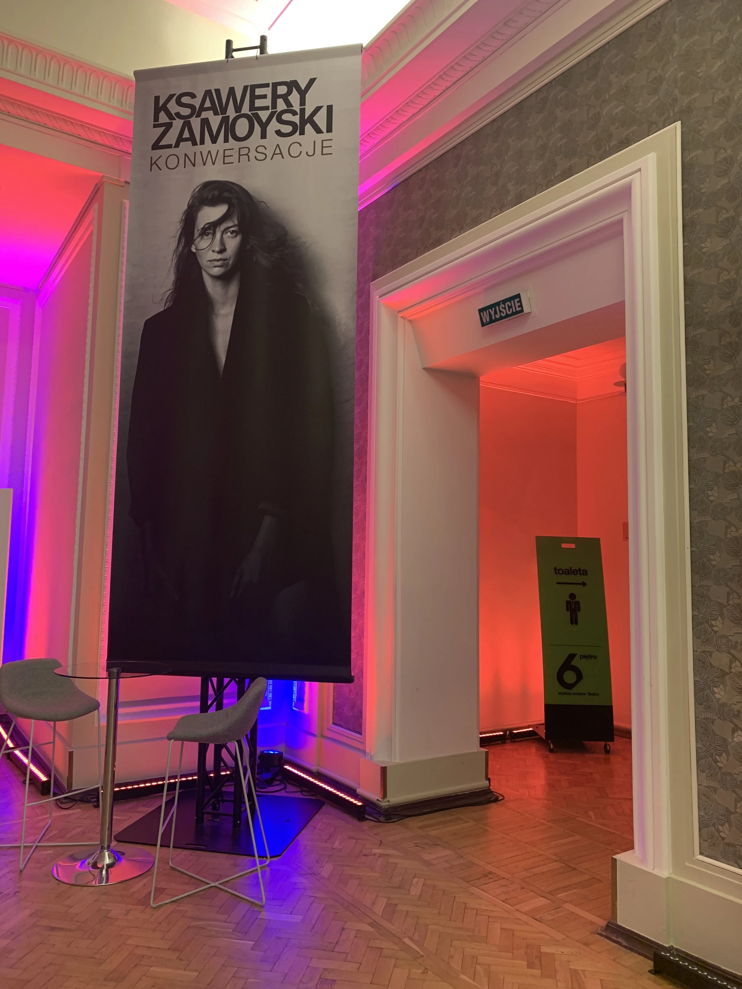 A black and white poster of Ksawery Zamoyski with the words 'Ksawery Zamoyski Konwersacje' at the top, hanging in a brightly lit room with pink and purple lighting. The room has ornate moldings and a parquet wood floor. There are two gray chairs and a small round table in front of the poster. An exit sign reads 'Wyjście' above a doorway leading to a dimly lit corridor with a green sign indicating restrooms and stairs.
