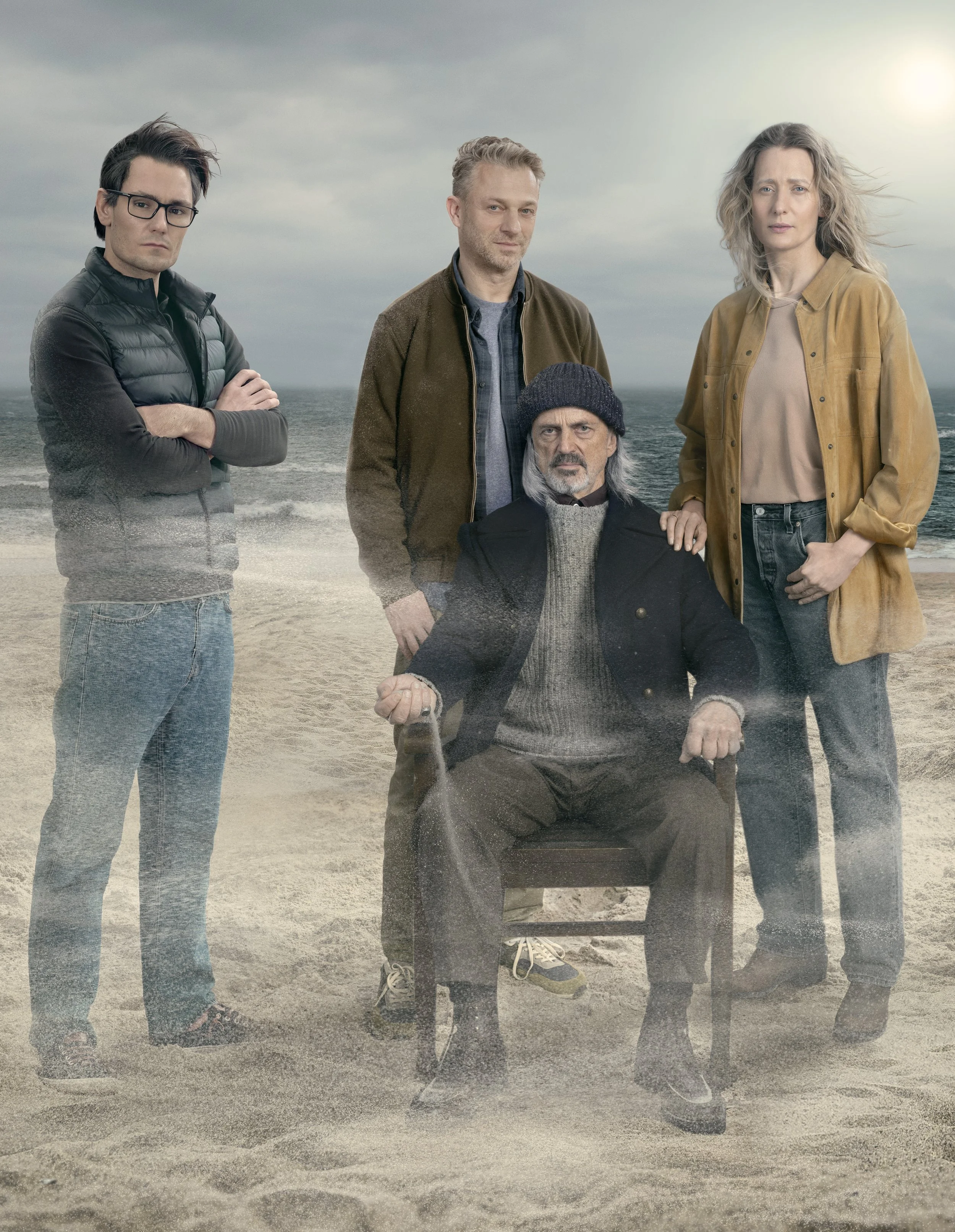 Four people standing on a beach with dark clouds overhead, one sitting in a chair, and the others standing beside him, all looking at the camera.