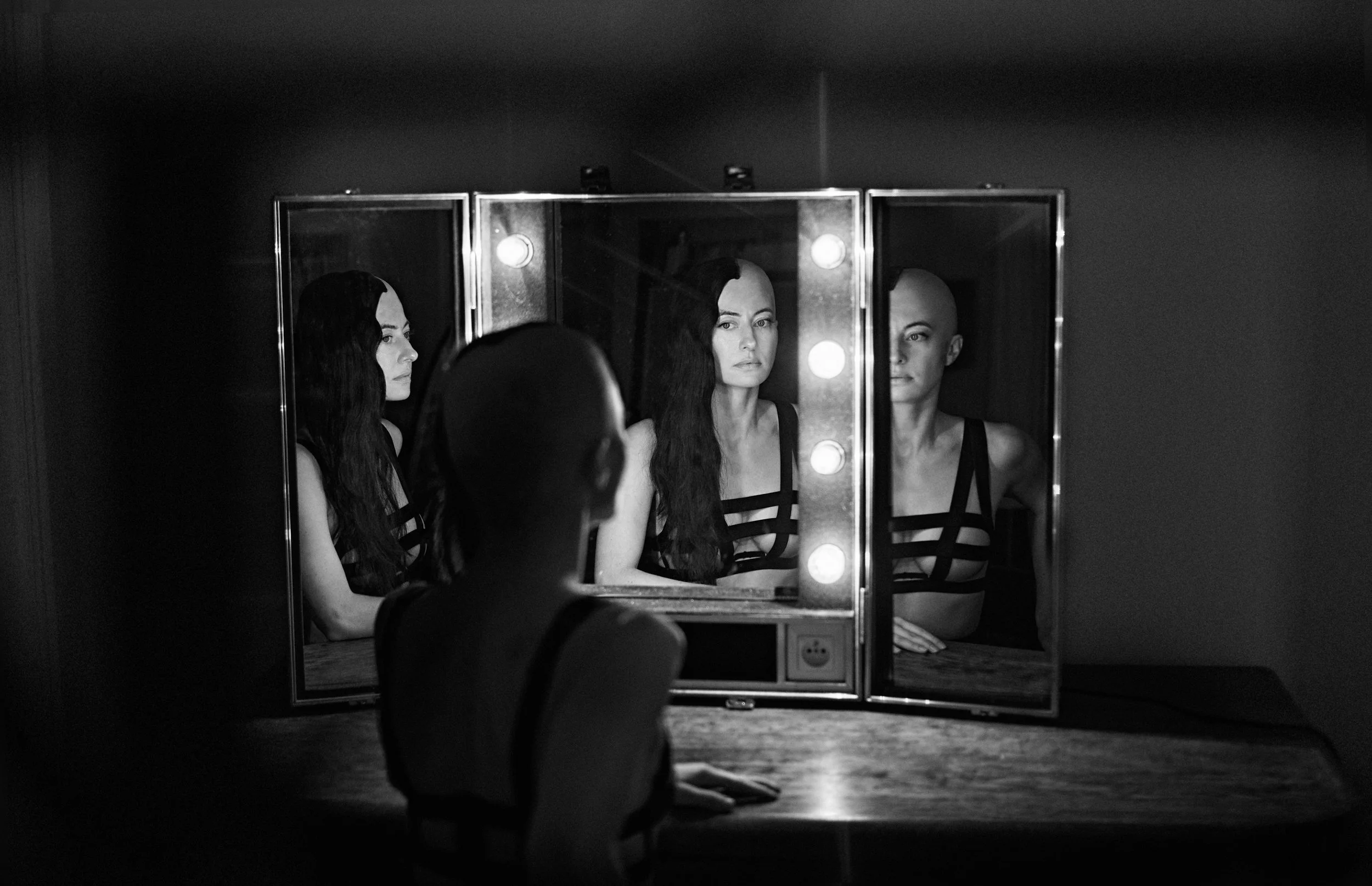 Black and white photo of a woman with long, wavy dark hair looking into a mirror with makeup bulbs, with similar images reflected in the mirror, including a woman who is bald and a woman with short hair, all wearing black clothing with straps.