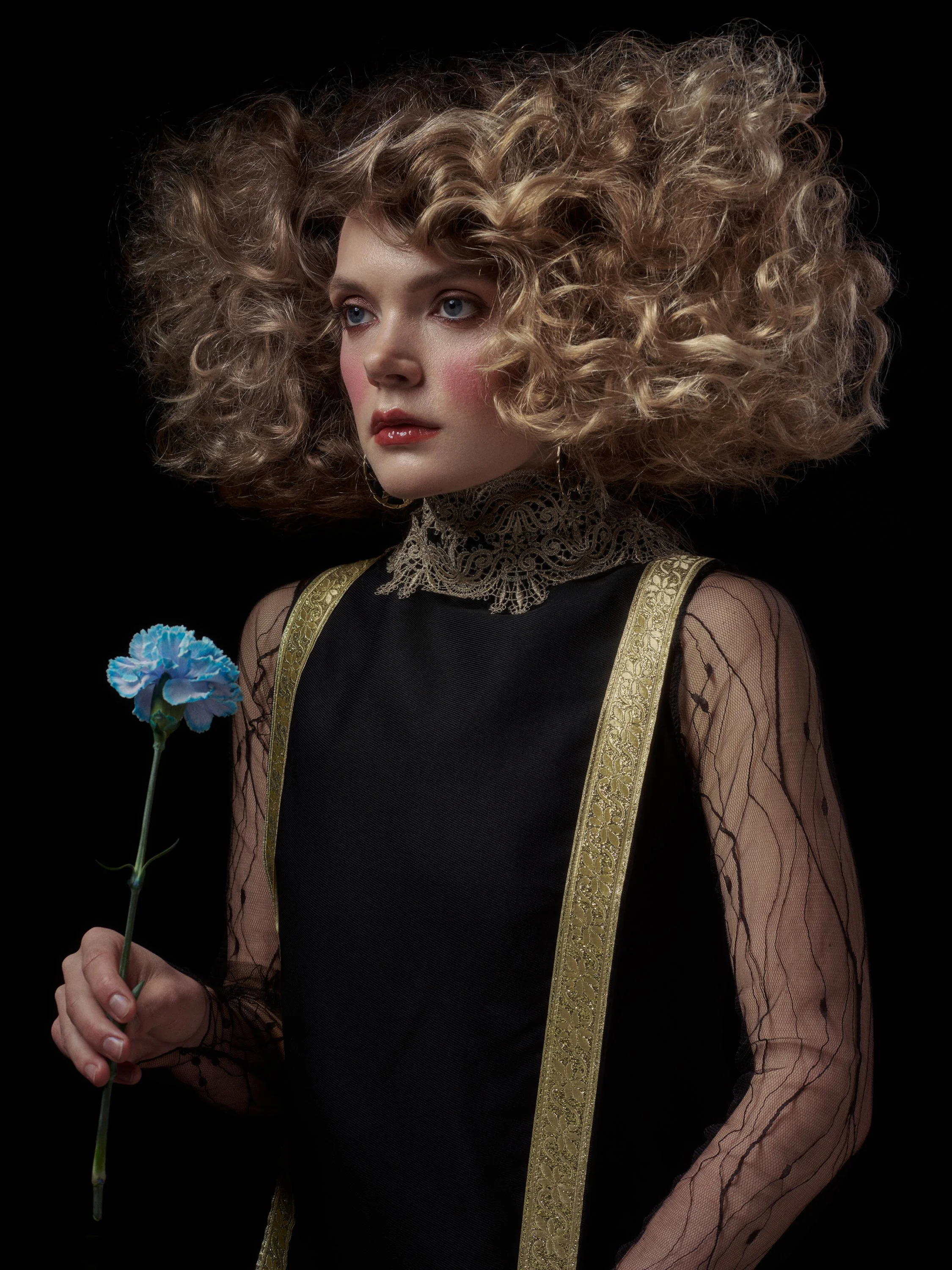 A woman with large, curly blonde hair against a black background, wearing a dress with lace and gold embroidered straps, holding a blue flower.