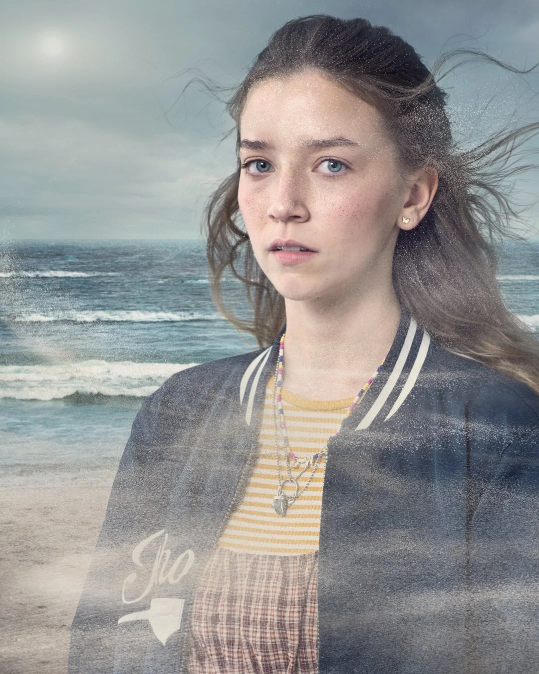 A young woman with blue eyes and long, wavy brown hair standing in front of the ocean on a cloudy day.