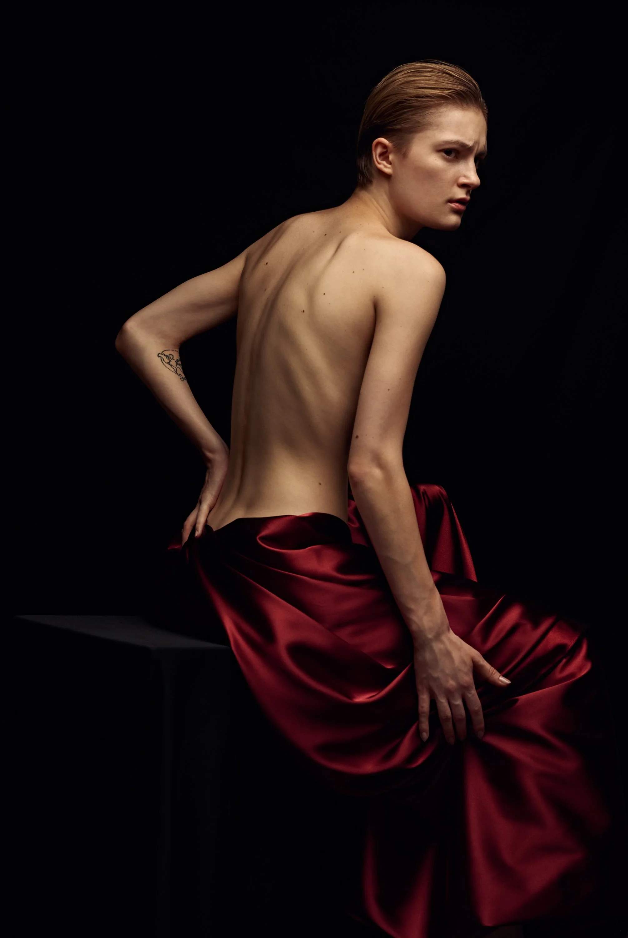 A topless woman with short blonde hair posed against a black background, wearing a red satin skirt, with one arm behind her back and the other resting on her thigh.