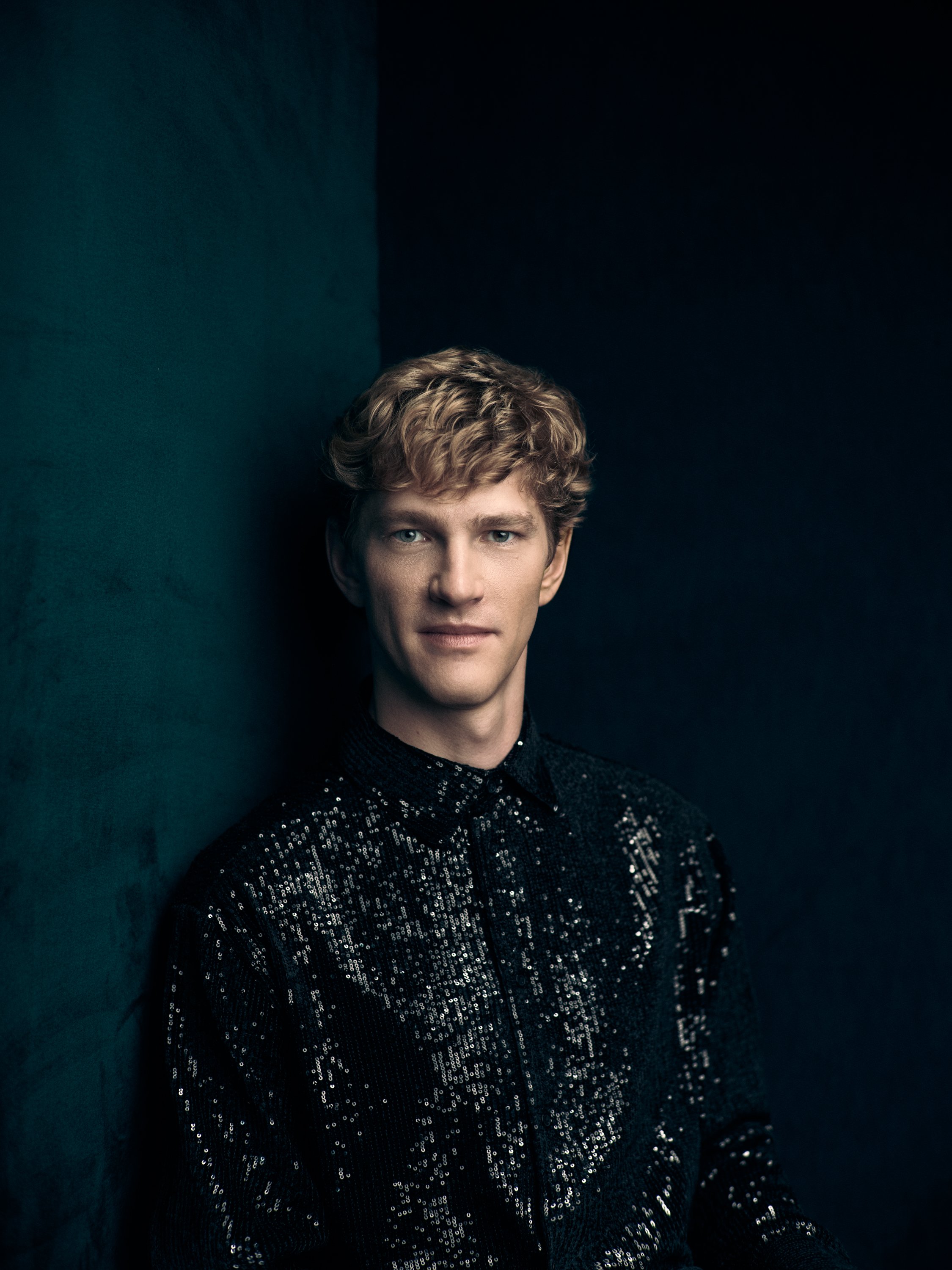 A young man with curly blond hair and blue eyes wearing a black sequined jacket, standing against a dark teal and black background. The portrait of Jan Lisiecki