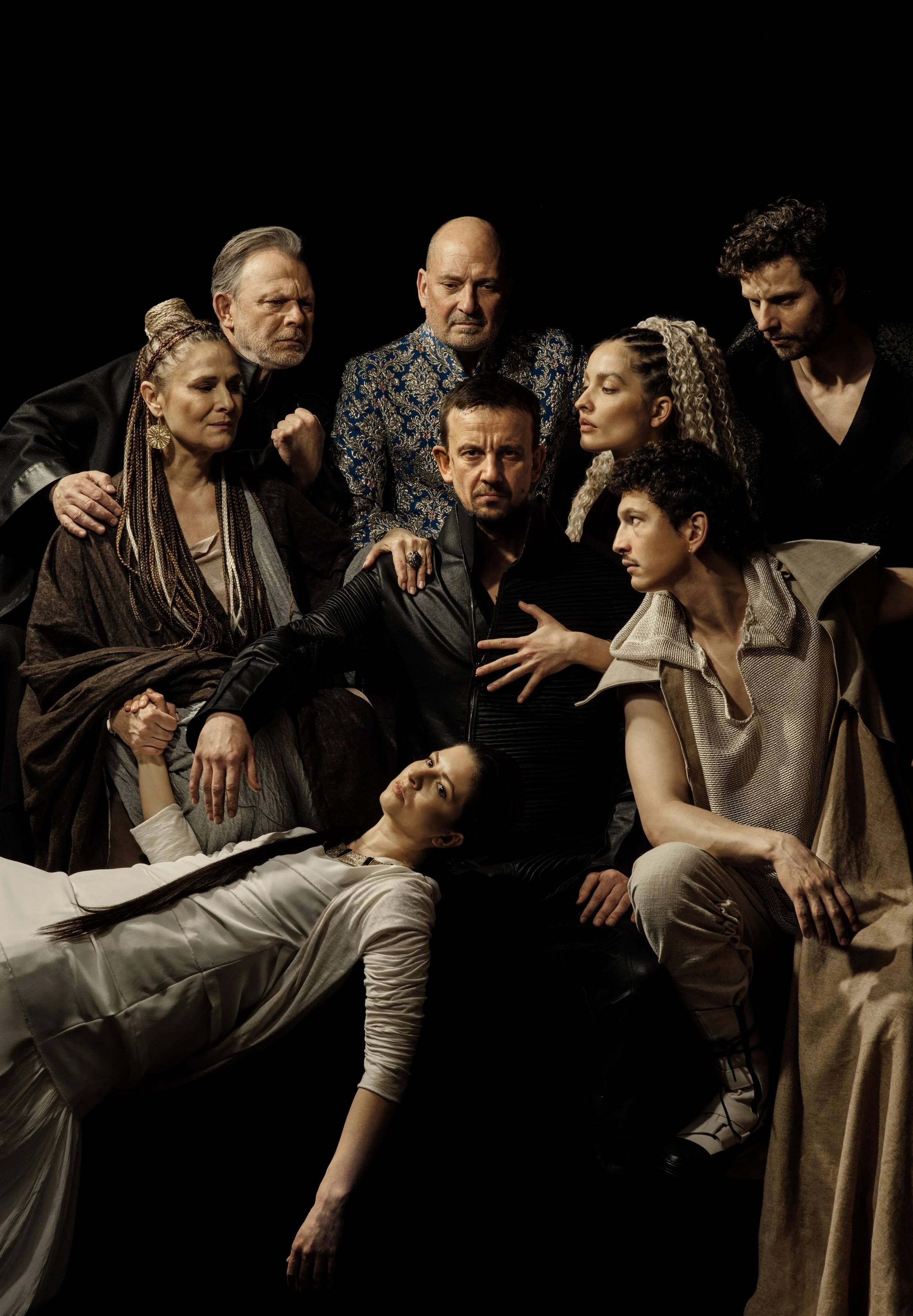 Group of nine diverse individuals in stylish clothing pose together against a black background, some in seated or reclined positions, with serious and contemplative expressions.