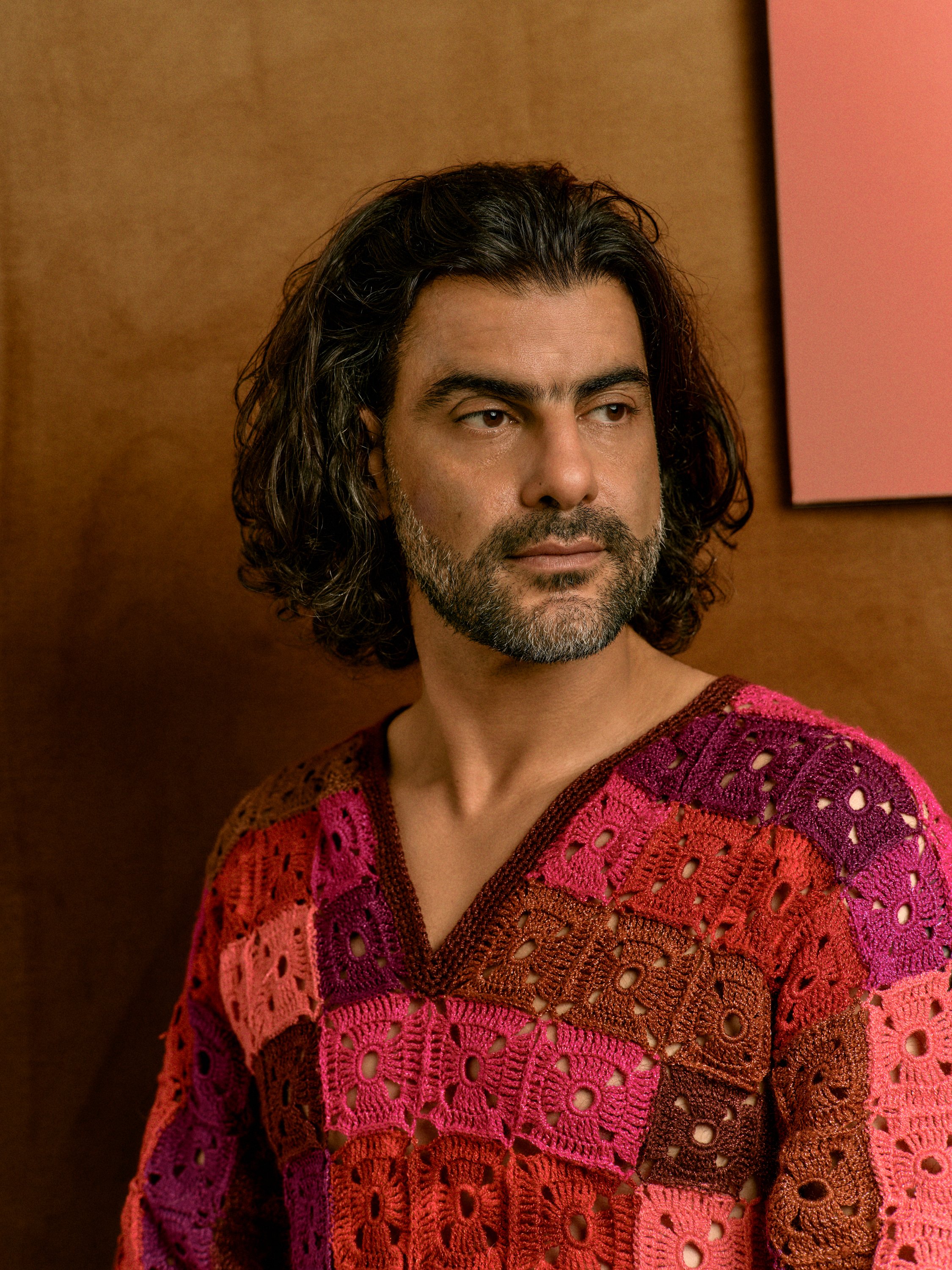A man with long, wavy dark hair and a gray beard, wearing a colorful crocheted sweater in shades of pink, red, orange, and purple, standing in front of a brown wall with a pink-framed picture. The portrait of Bassem Akiki