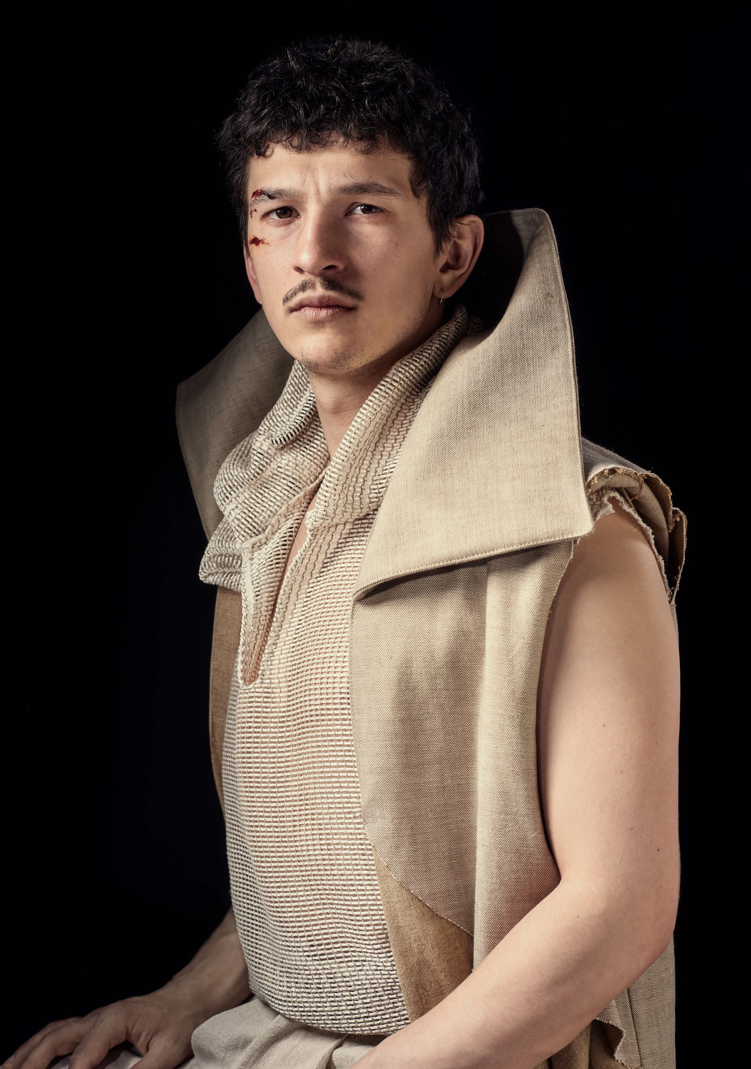 A man with short dark hair and a mustache, looking directly at the camera, has a bruise and cut on his left cheek. He wears a sleeveless beige coat with a high collar and a beige checked shirt underneath. The background is black.