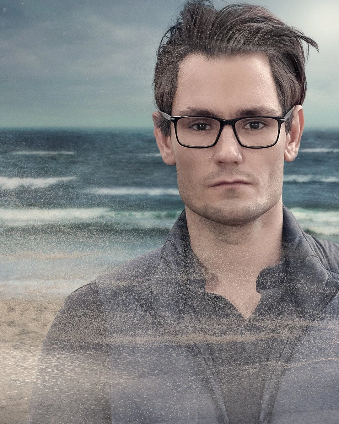 A young man with dark hair, glasses, and a serious expression, standing on a beach with waves and an overcast sky in the background.