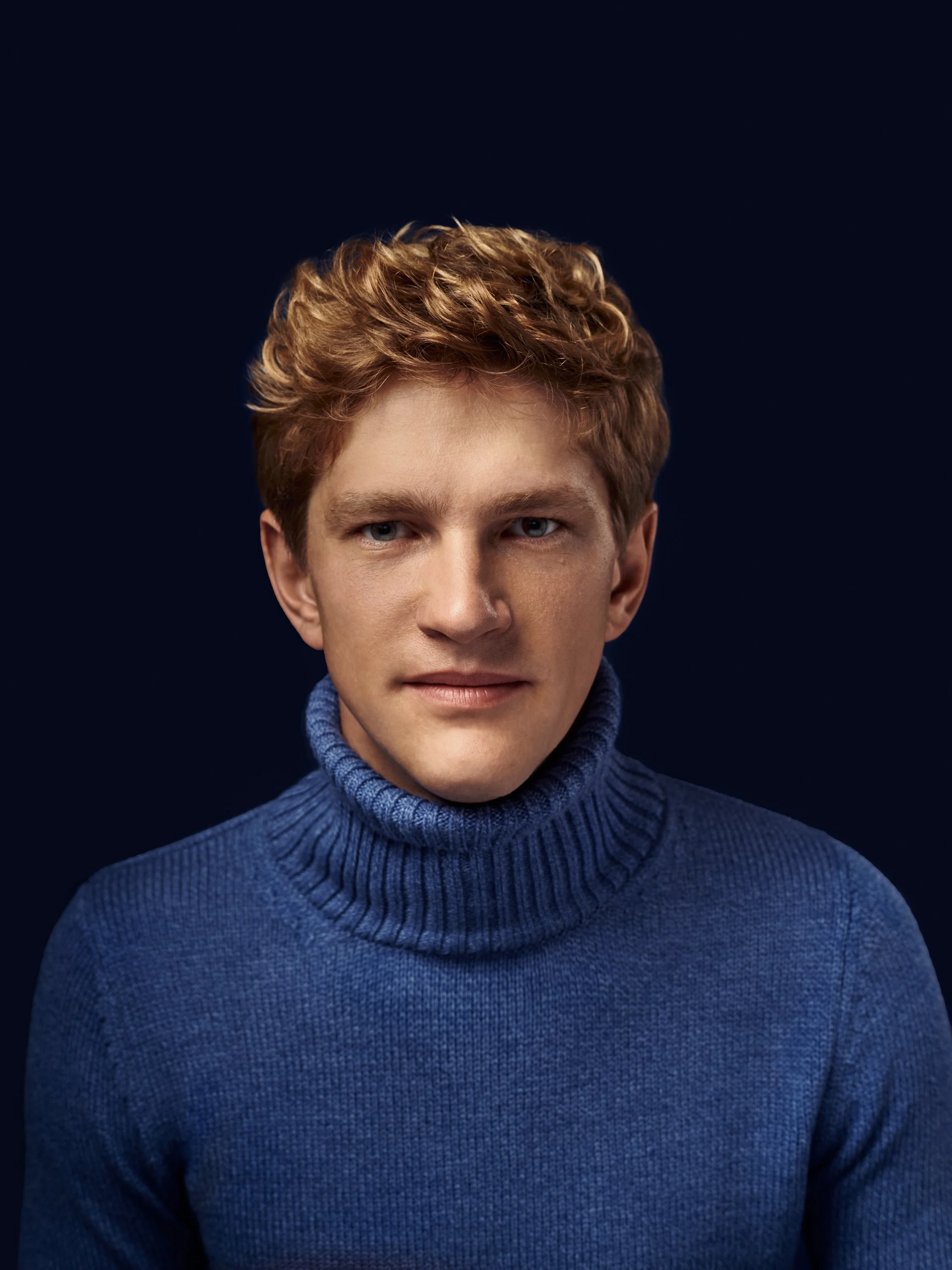 Close-up of a young man's face with short, curly red hair, blue eyes, and fair skin, wearing a knitted blue turtleneck sweater, against a dark background.