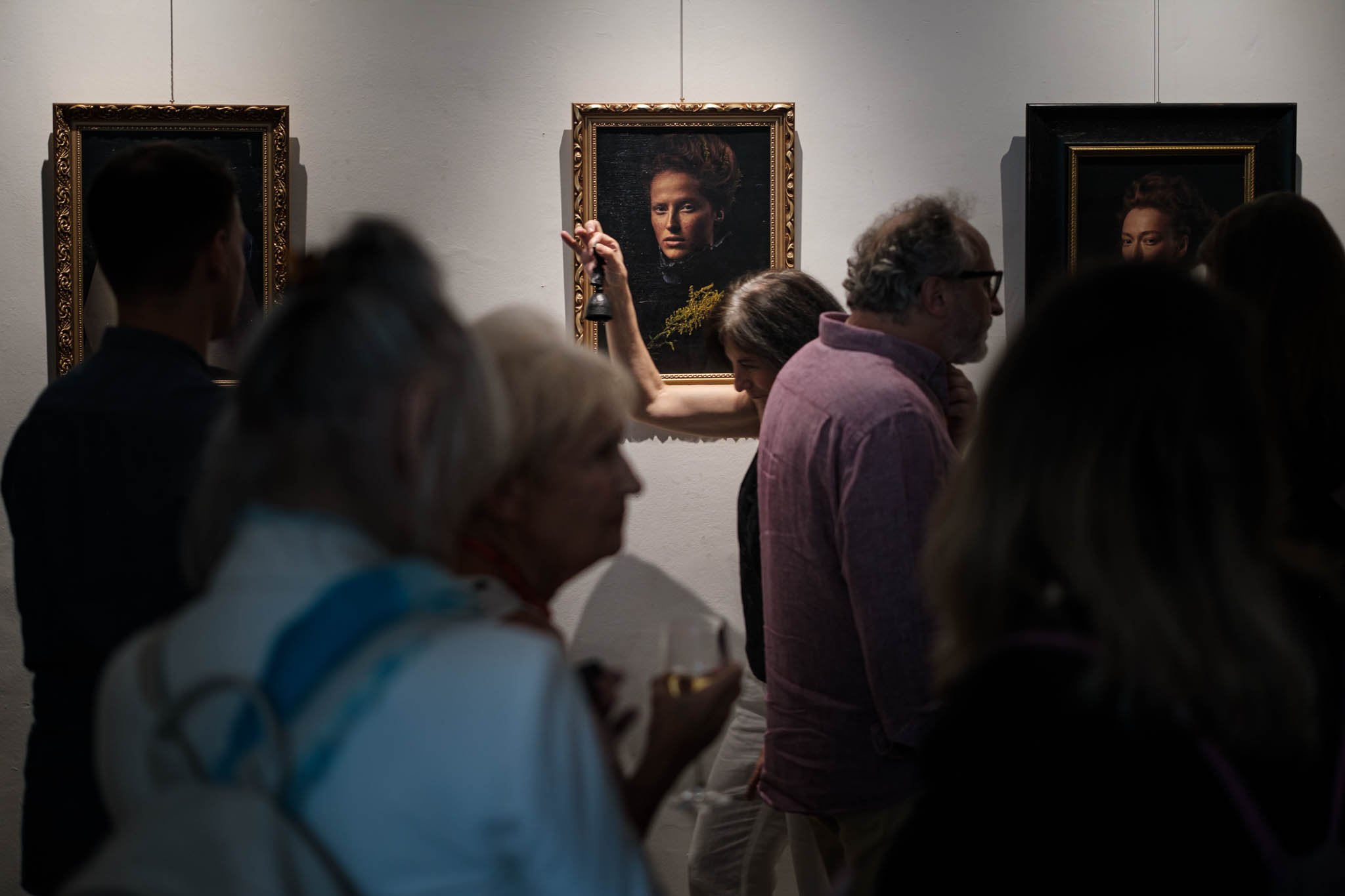 People attending an art gallery, observing framed portraits on a white wall, with one person raising an arm, possibly taking a photo or pointing, in a dimly lit room.