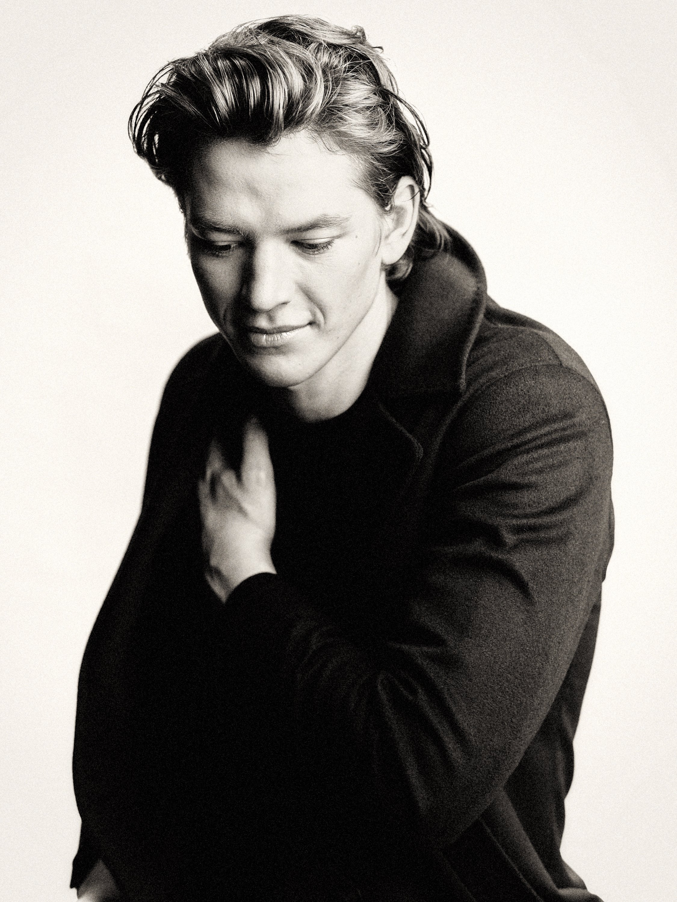 Black and white photo of a young man with styled hair, looking down and smiling slightly, wearing a dark jacket.