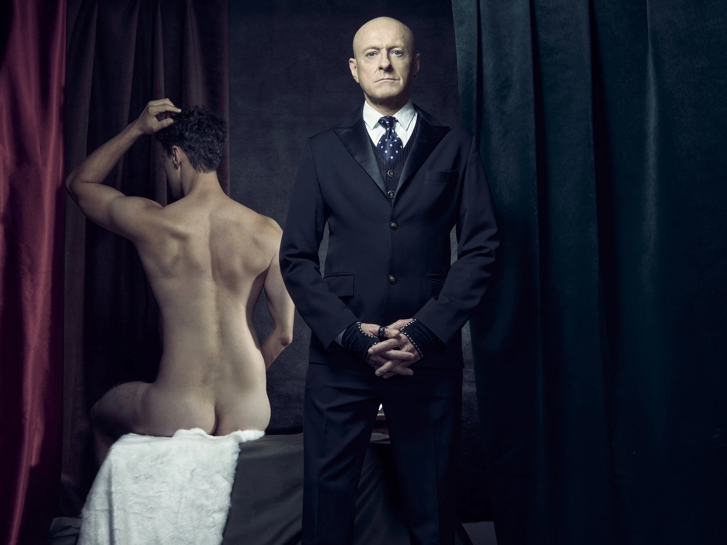 A large, bald man in a dark suit standing next to a topless person with short curly hair, who appears to be adjusting their hair. They are on a stage with dark curtains around them.