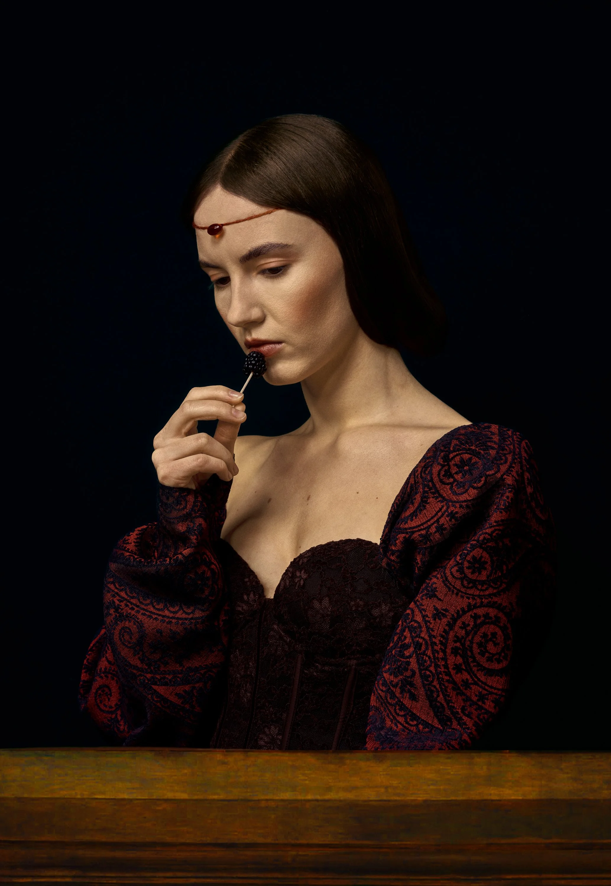 A woman with dark brown hair, wearing a red and black patterned dress, sits at a wooden table with her chin resting on her hand, looking thoughtful, holding a blackberry on a toothpick near her lips, against a dark background. Martyna Byczkowska