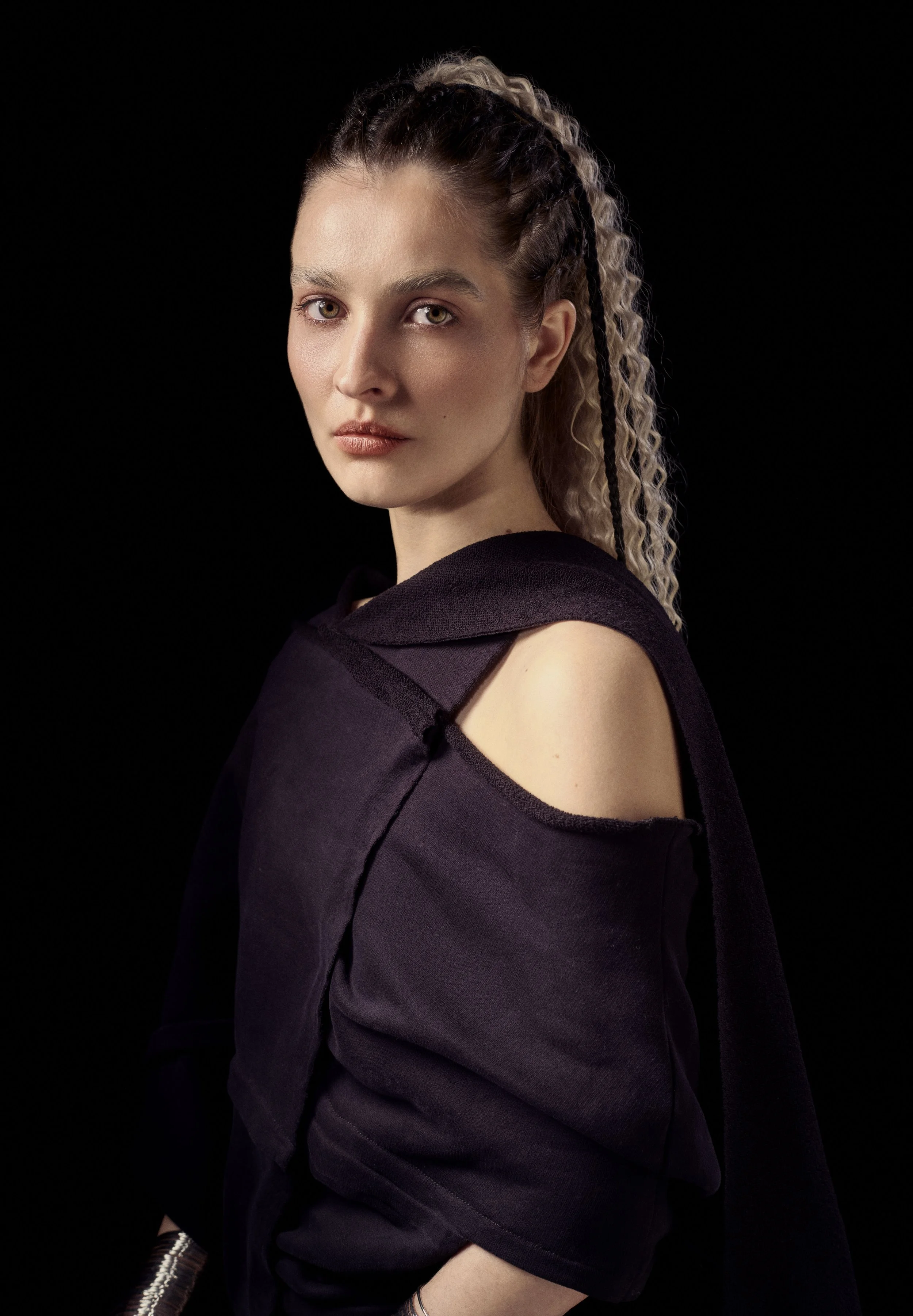 A woman with light skin tone, light-colored eyes, and curly hair styled in braids, wearing a dark off-shoulder top, against a black background.