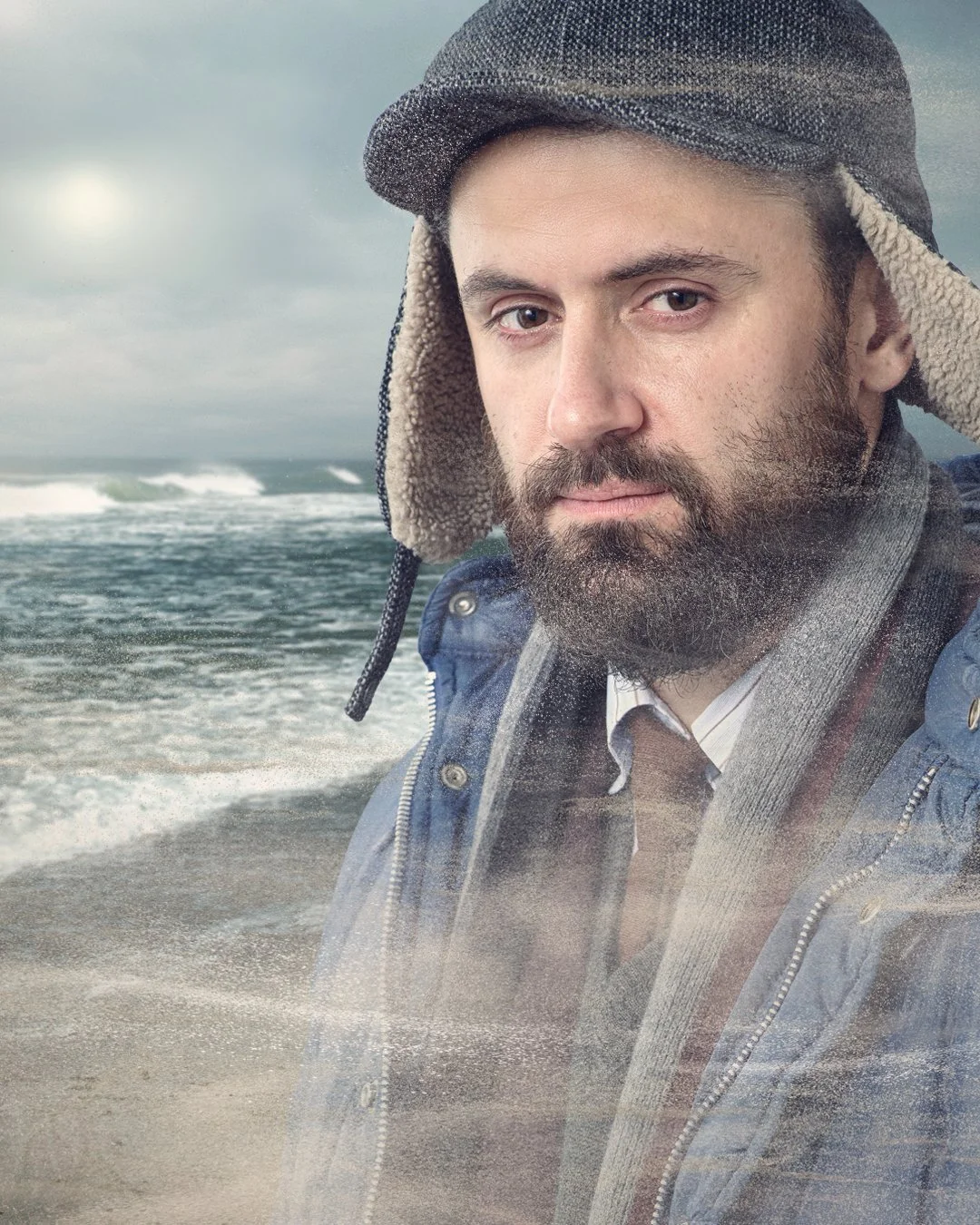 A man with a beard wearing a warm hat and jacket stands on a beach during overcast weather, with ocean waves in the background.