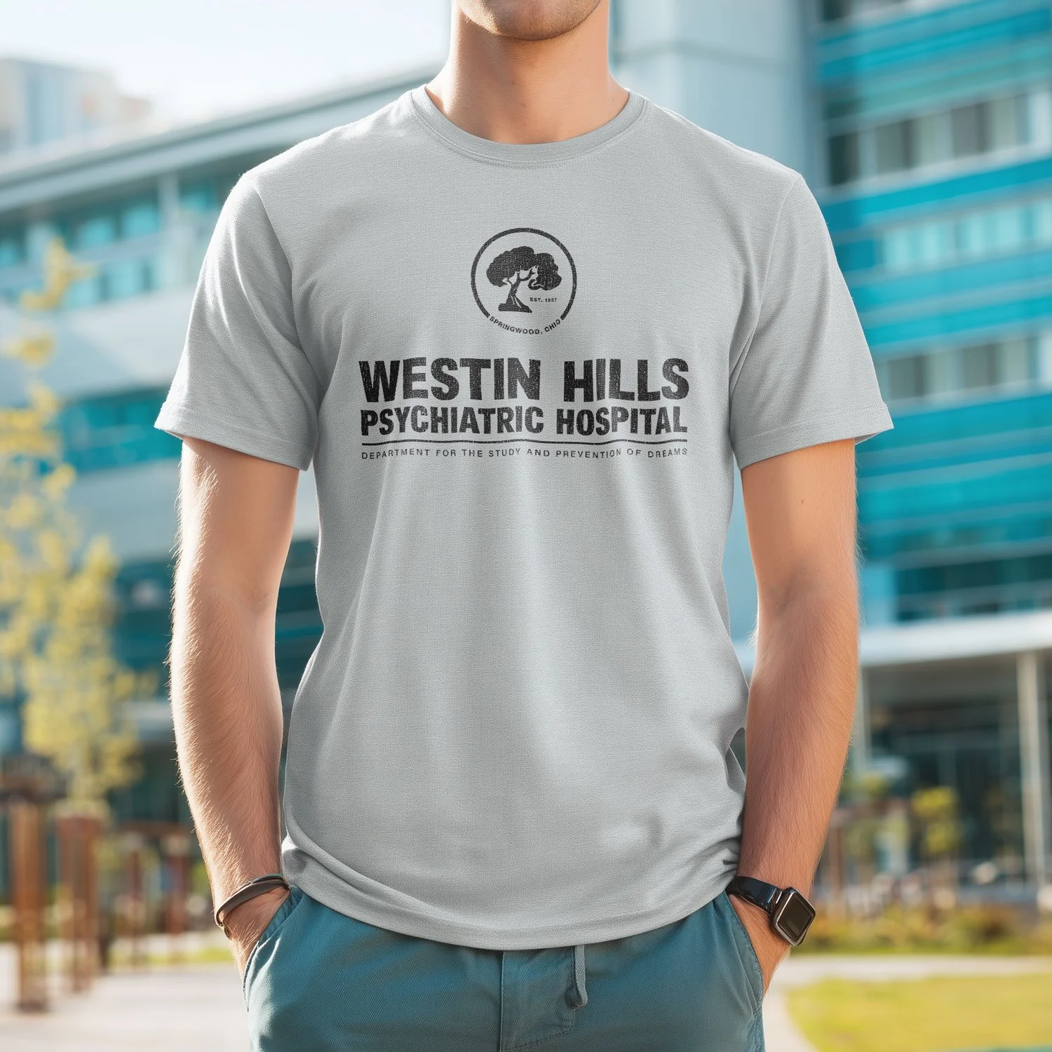 A man wearing a Sport Grey Westin Hills Psychiatric Hospital T-shirt stands outside a hospital with a creepy atmosphere. The shirt was inspired by the movie A Nightmare on Elm Street.