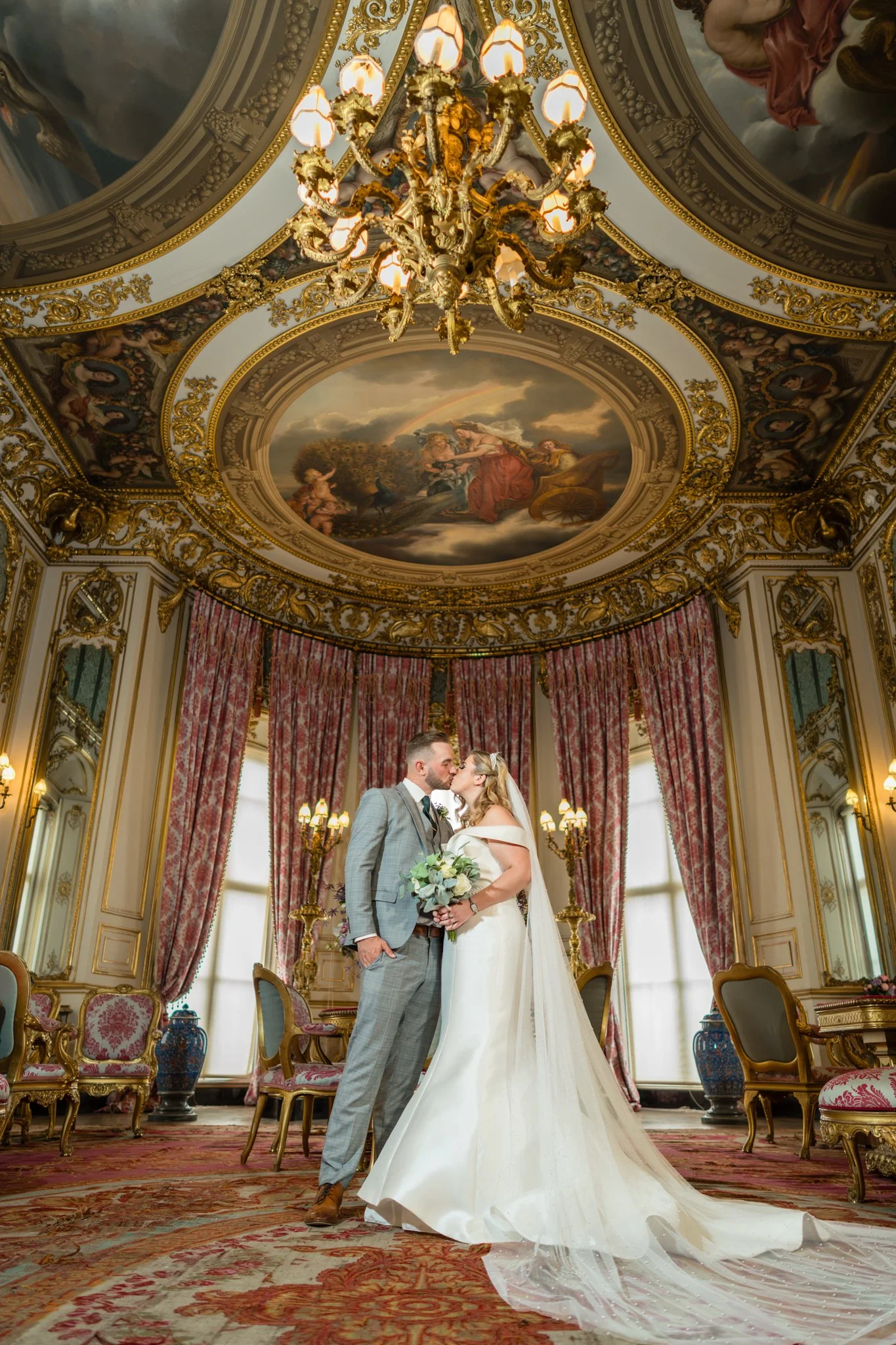 Bride and groom sharing a romantic kiss in the opulent State Room at Belvoir Castle, Leicestershire – gilded dome ceiling, red damask curtains, chandeliers, and luxurious Regency interiors as a perfect fairytale wedding setting