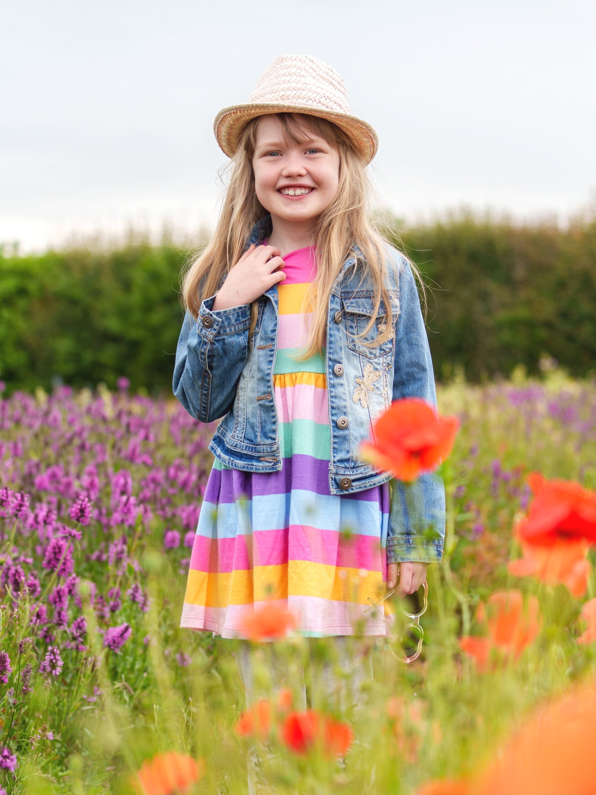 Image of child wearing a brightly coloured striped dress, denim jacket and straw hat standing in a field surrounded by wild flowers.