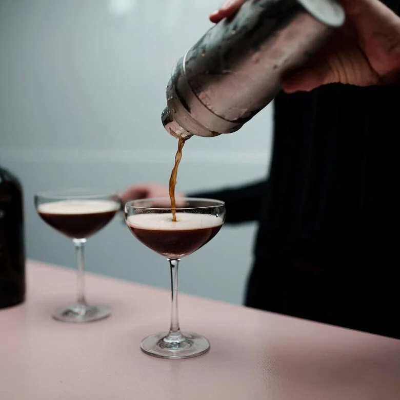 A person pouring a dark cocktail from a shaker into a coupe glass, with another similar cocktail in the background.