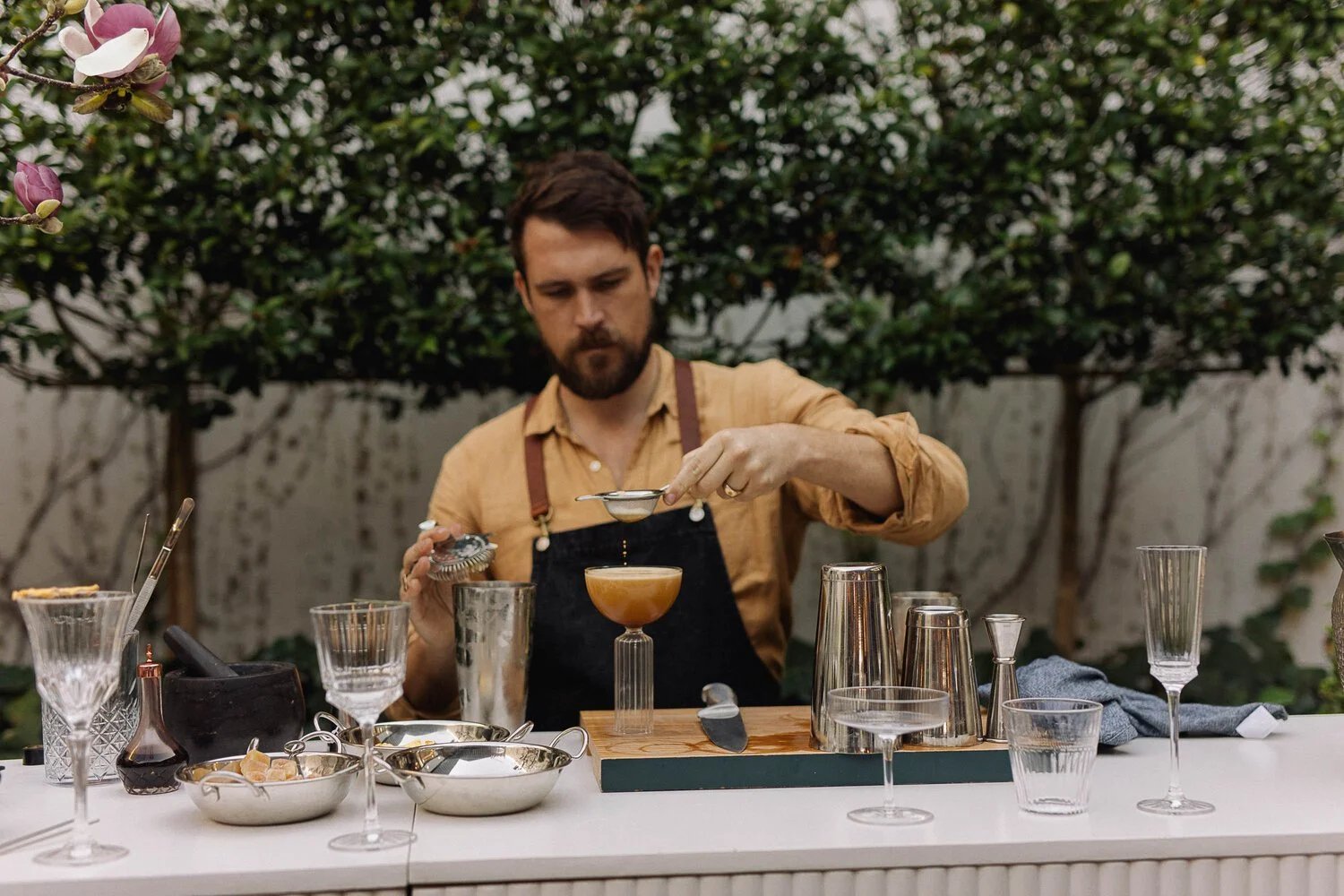 Man preparing a cocktail outdoors with various bar tools and glasses on a white table, greenery in the background.