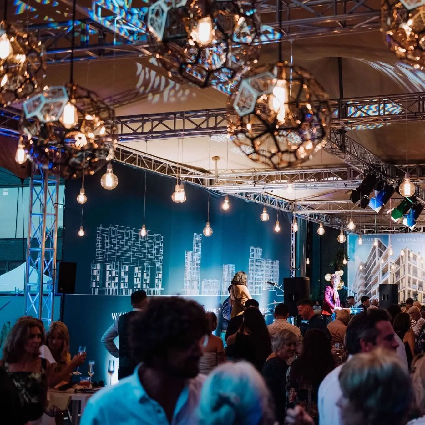 Indoor event with a stage, hanging lights, and large crowd of people, some seated and some standing, with a backdrop displaying a city skyline.