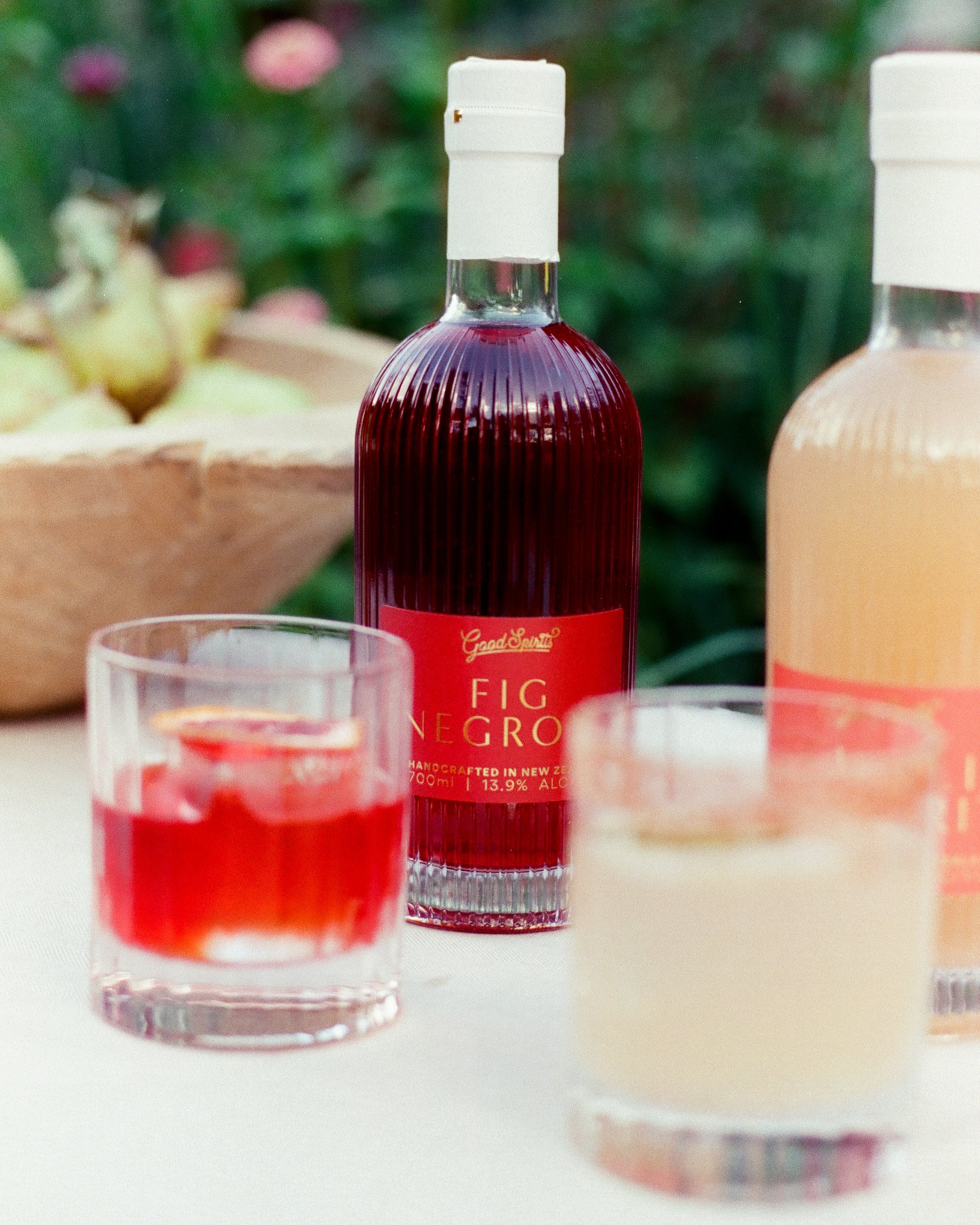 A bottle of Fig Negro liquor with two glasses, one filled with red liquid and one with a pale drink, on a white table with a bowl of green grapes in the background.
