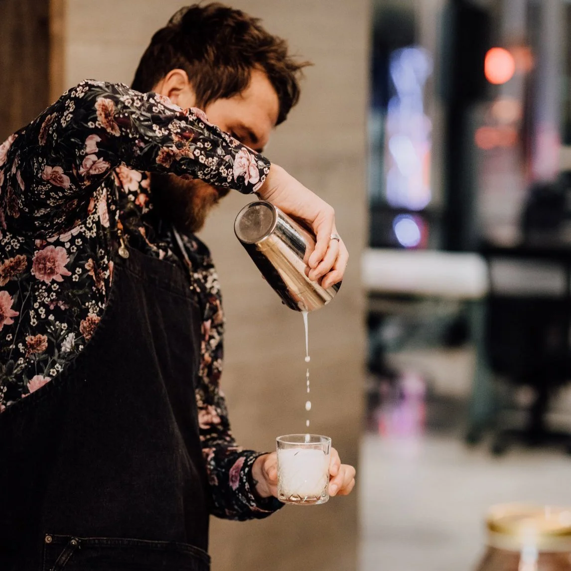 A man with a floral shirt and black apron pouring a drink from a shaker into a glass.
