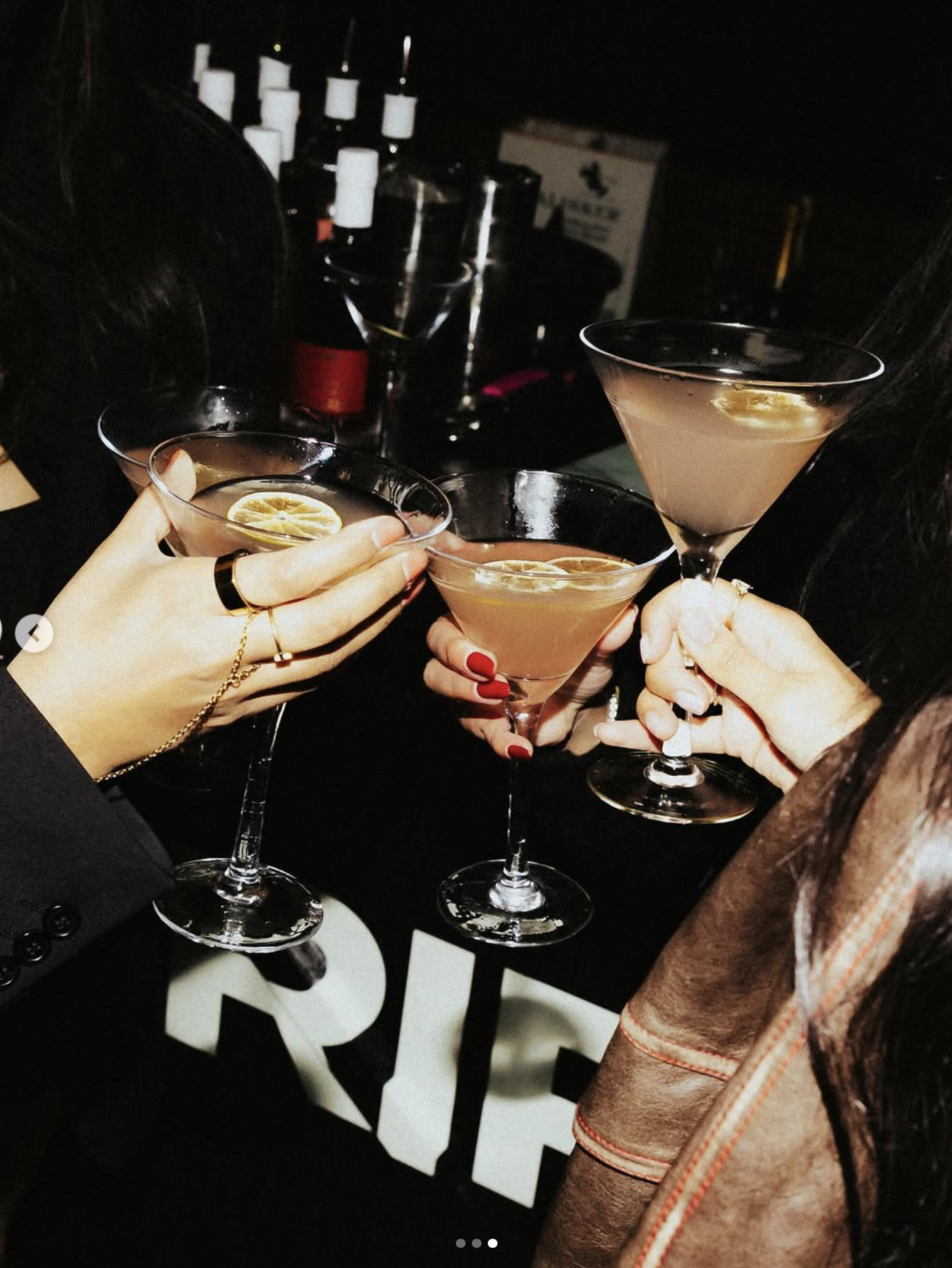 Three women clinking martini glasses with lemon slices in a dark bar setting.