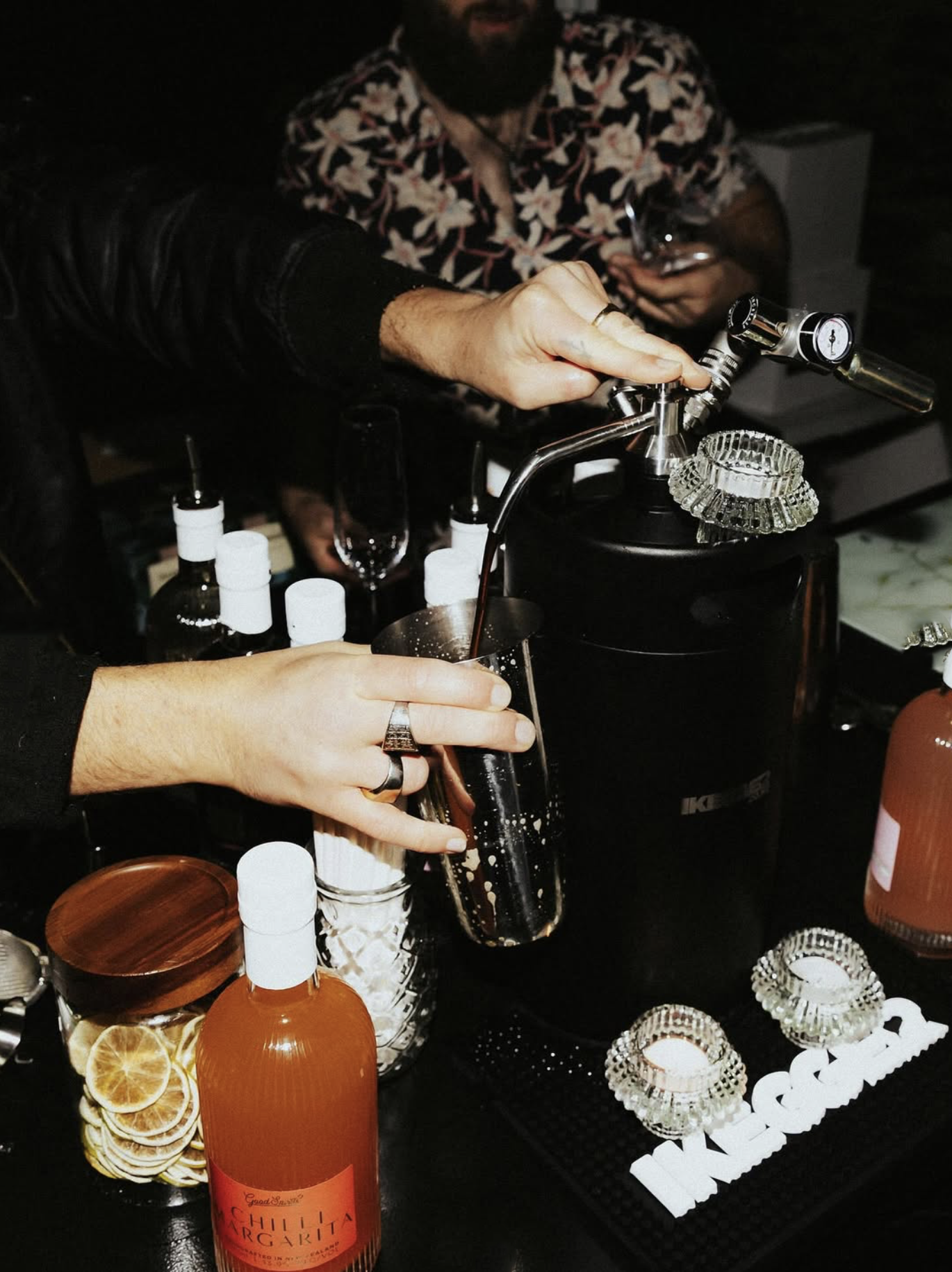 A person is pouring a dark beverage from a siphon or dispenser into a glass, with another person holding a wine glass in the background, surrounded by bottles, candles, and decorative items on a dark table.