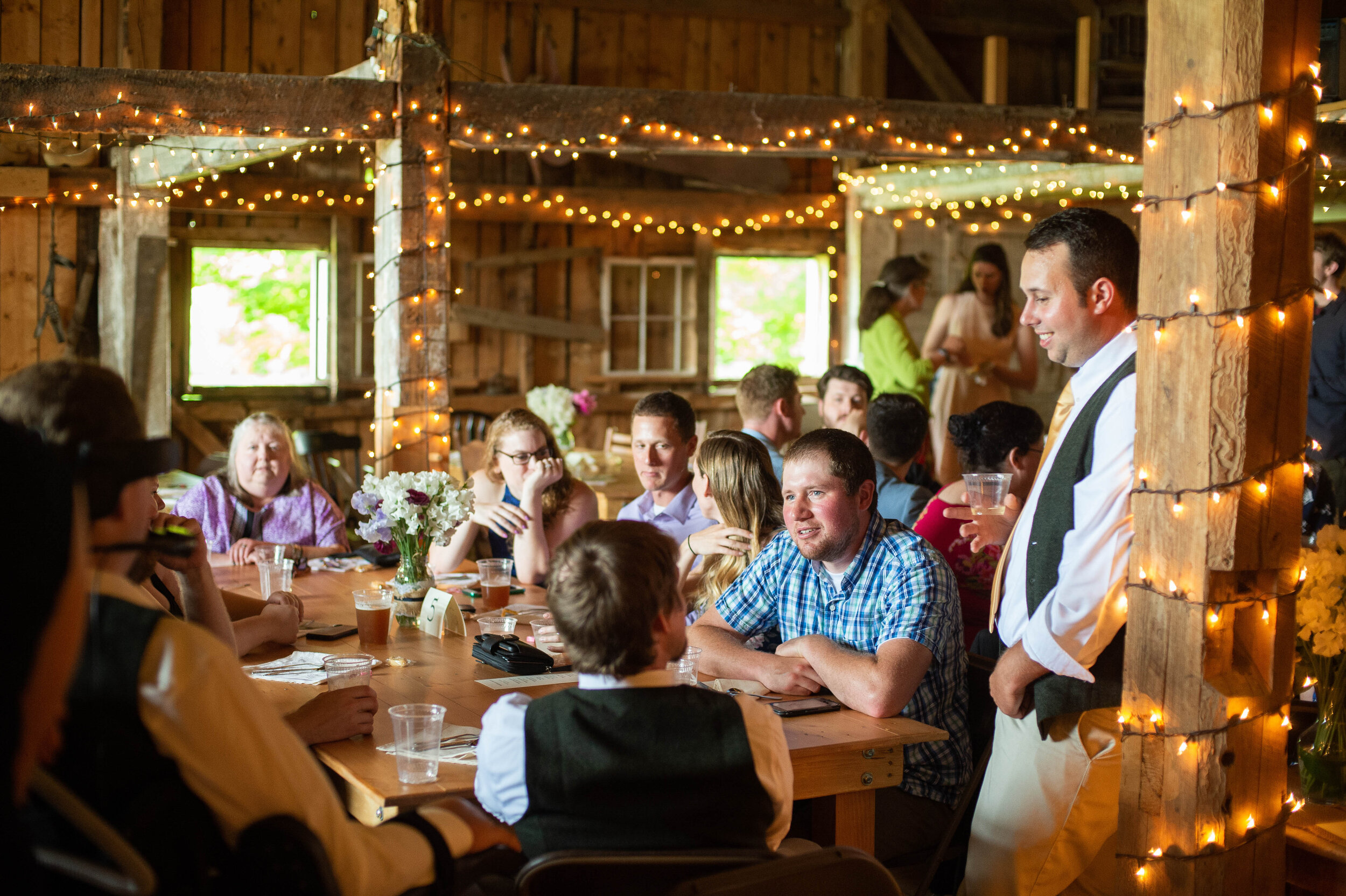 A gathering of people seated in a barn lit with twinkle lights in daylight