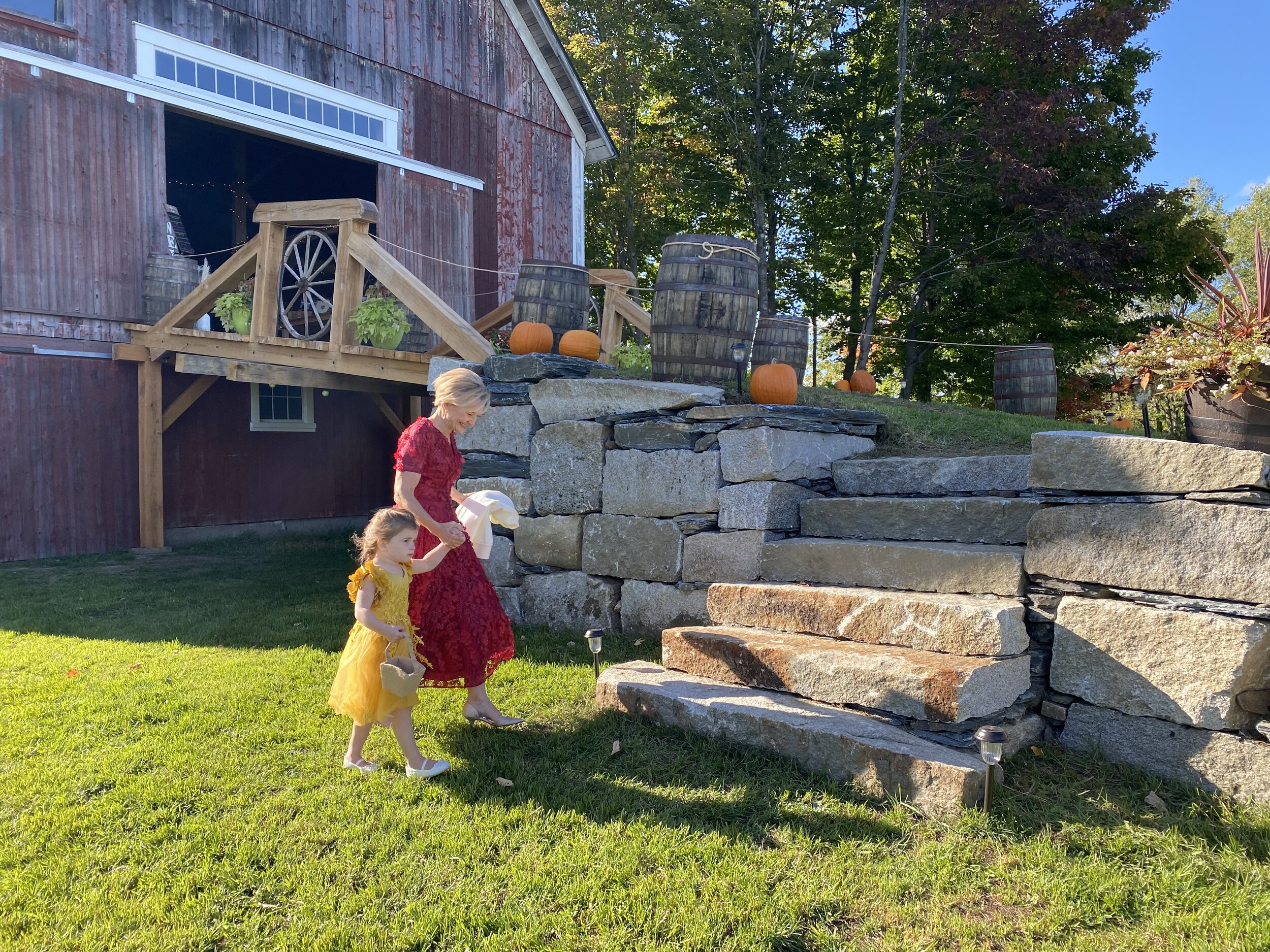 A woman and child dressed up walking up to a rustic barn with pumpkins placed outside