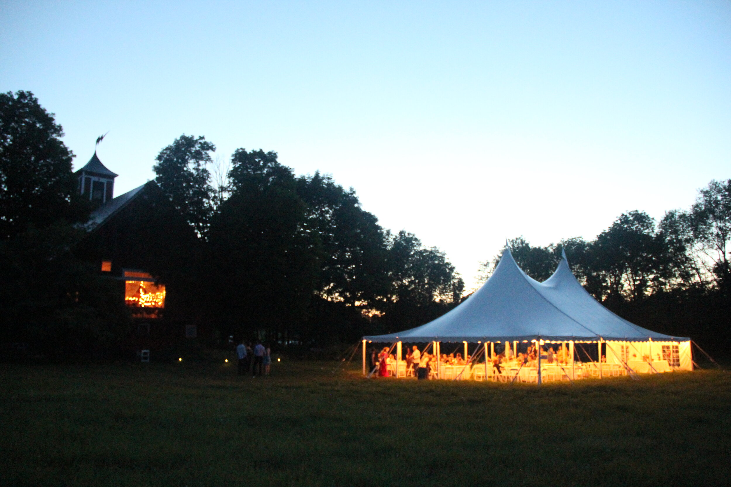 A wedding tent lit up at dusk outside of a barn