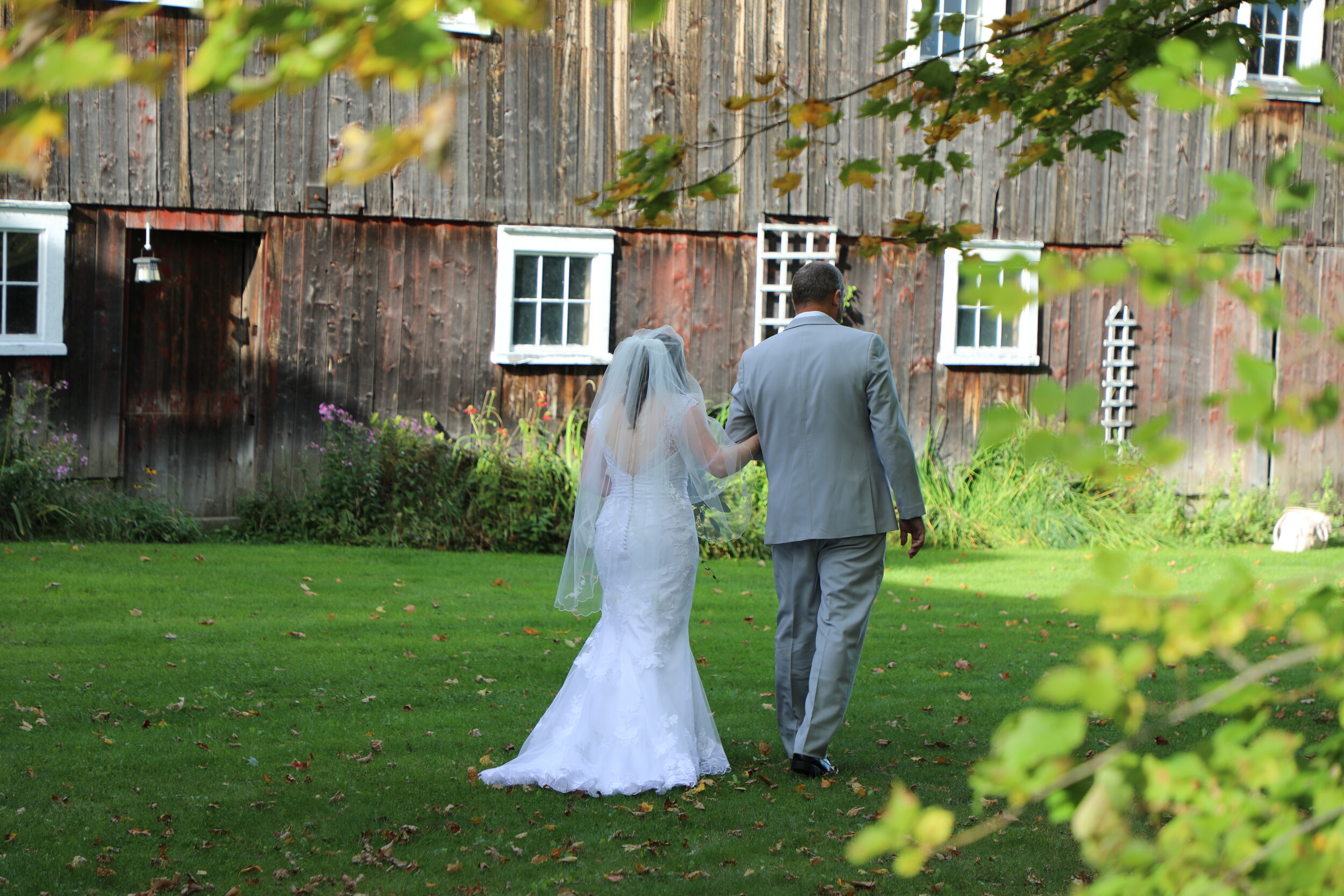 A bride and groom walking towards a rustic barn in the summer