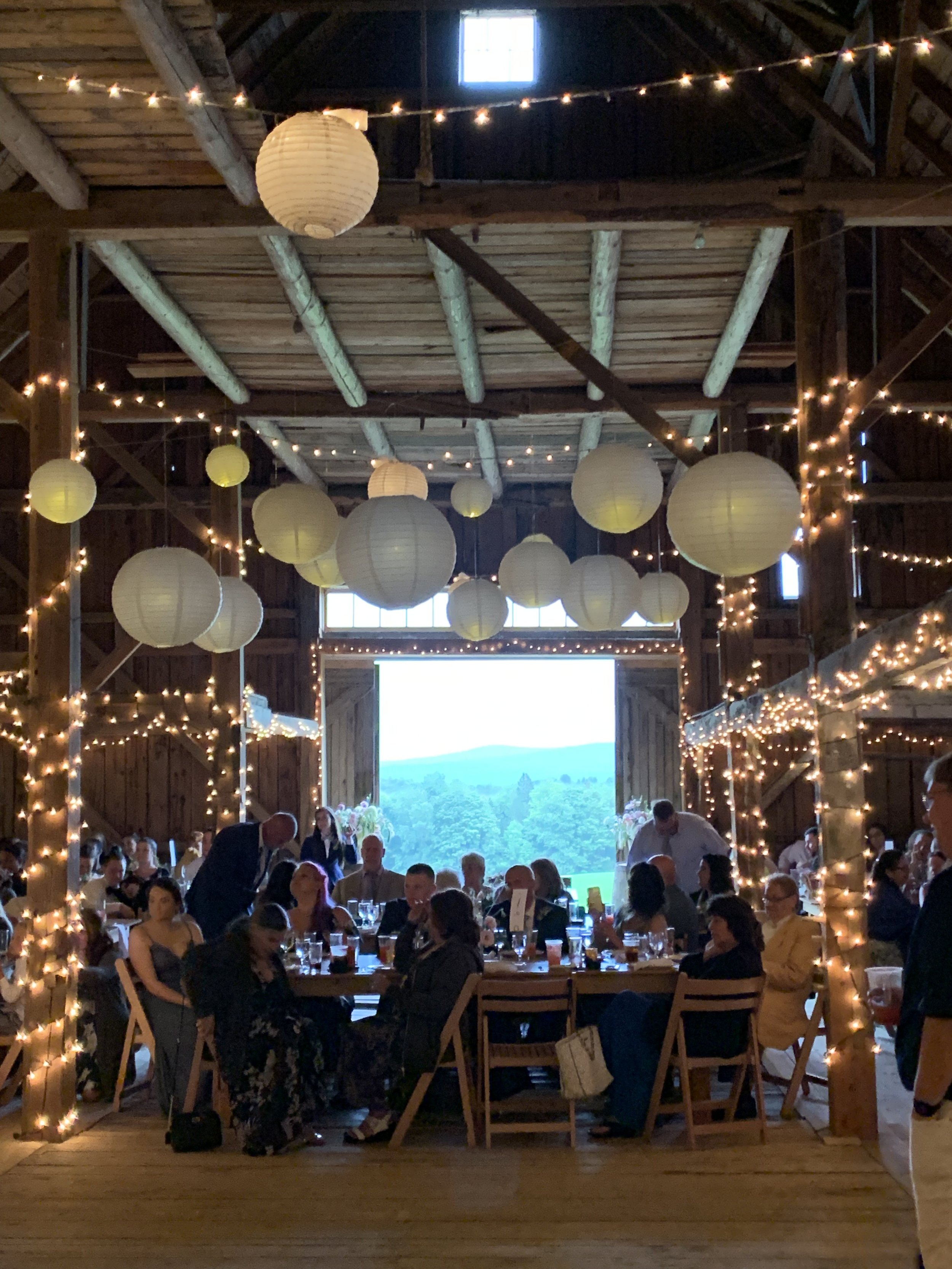Wedding guests in a lit barn at tables with a view of Vermont hills