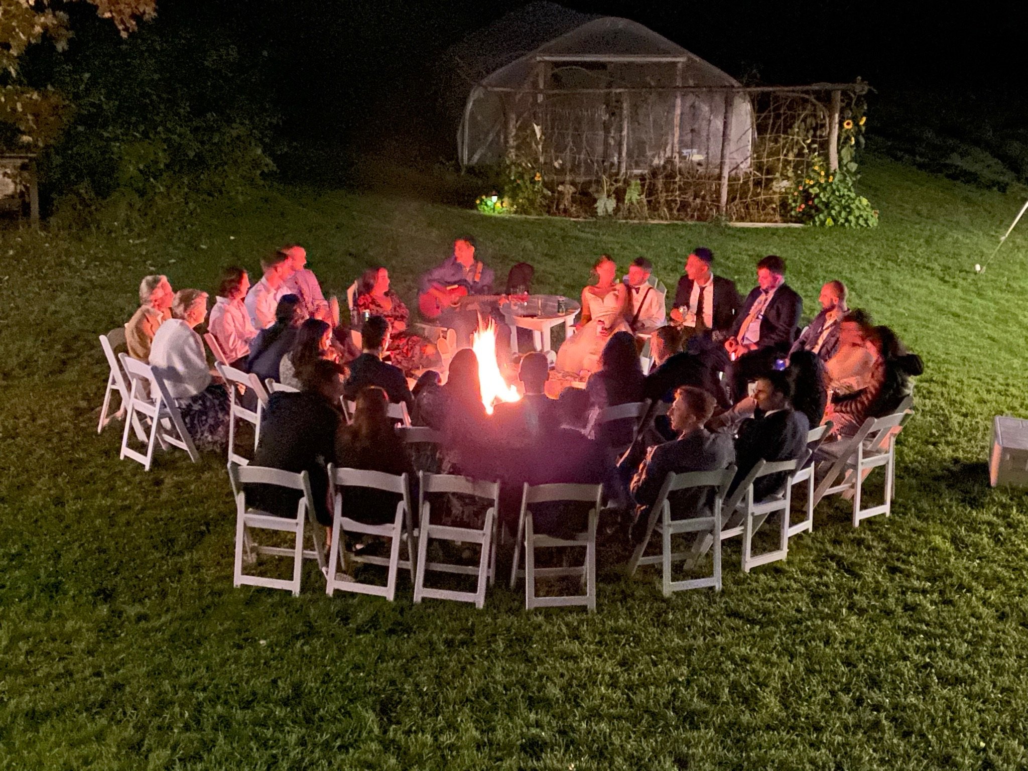 A bride and groom and their guests seated around a campfire outside the barn
