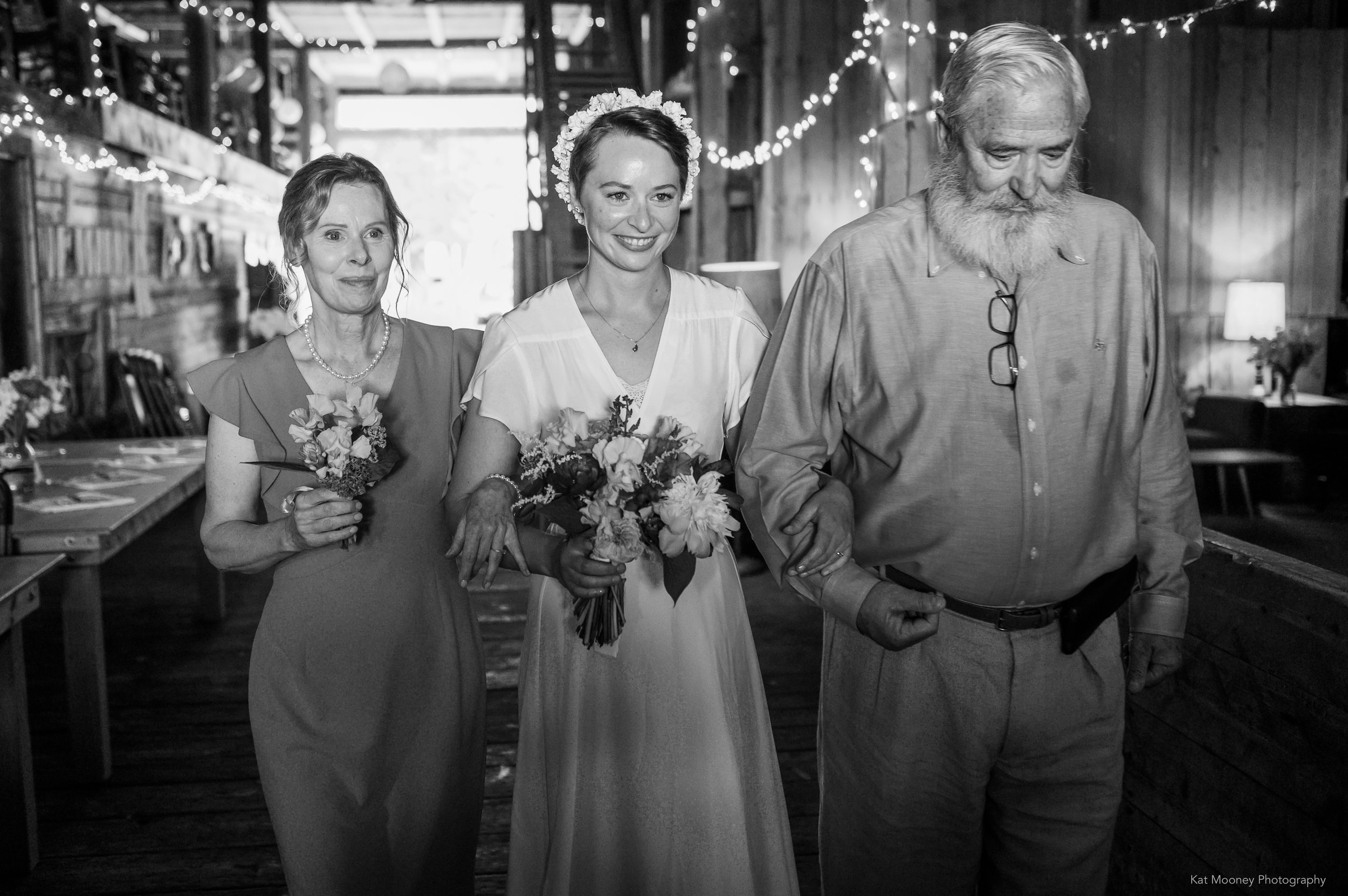 A bride surrounded by her parents headed to the aisle inside of a decorated barn
