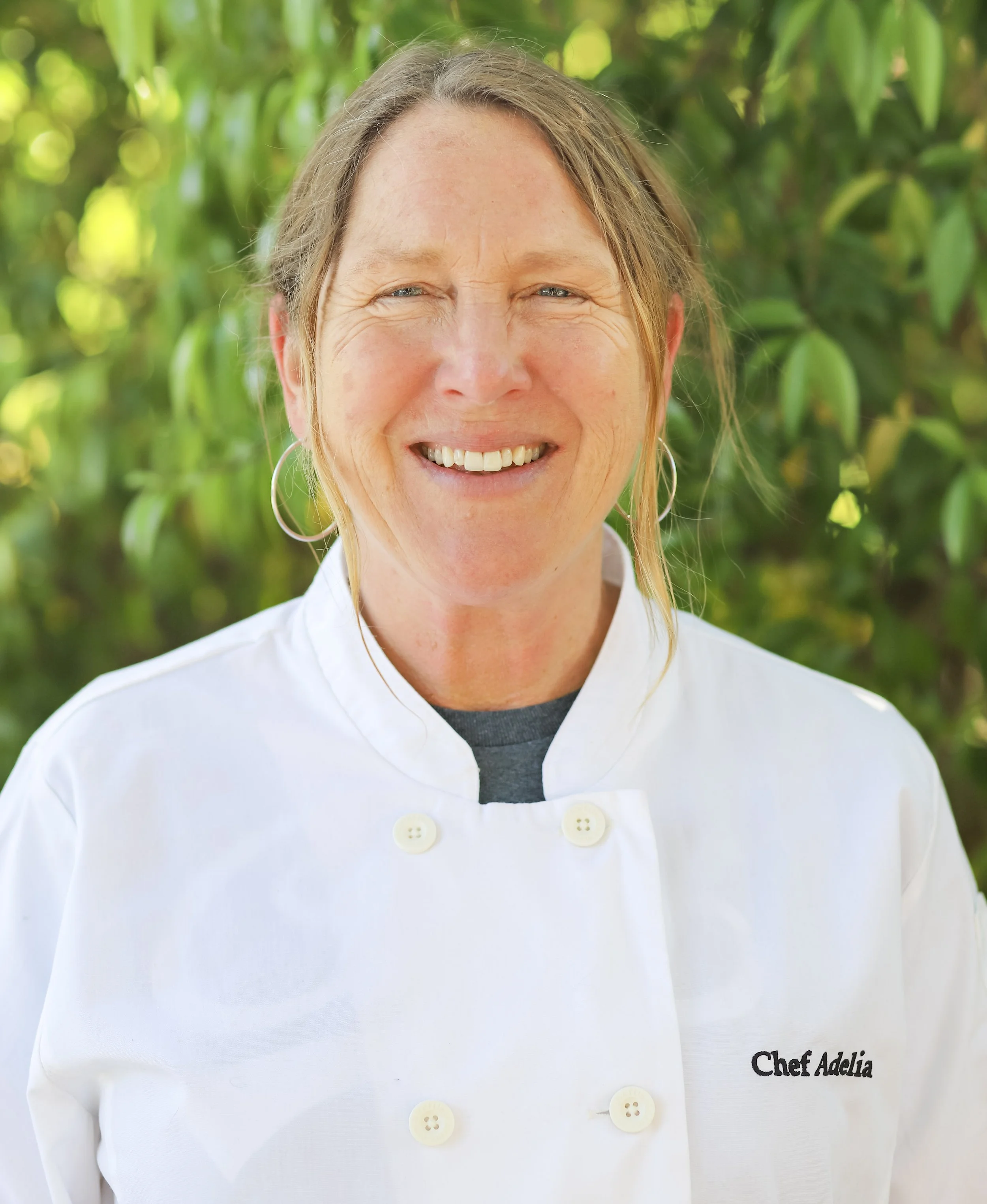 Smiling woman in a white chef's coat with "Chef Adelia" embroidered on it, outdoors with greenery in the background.