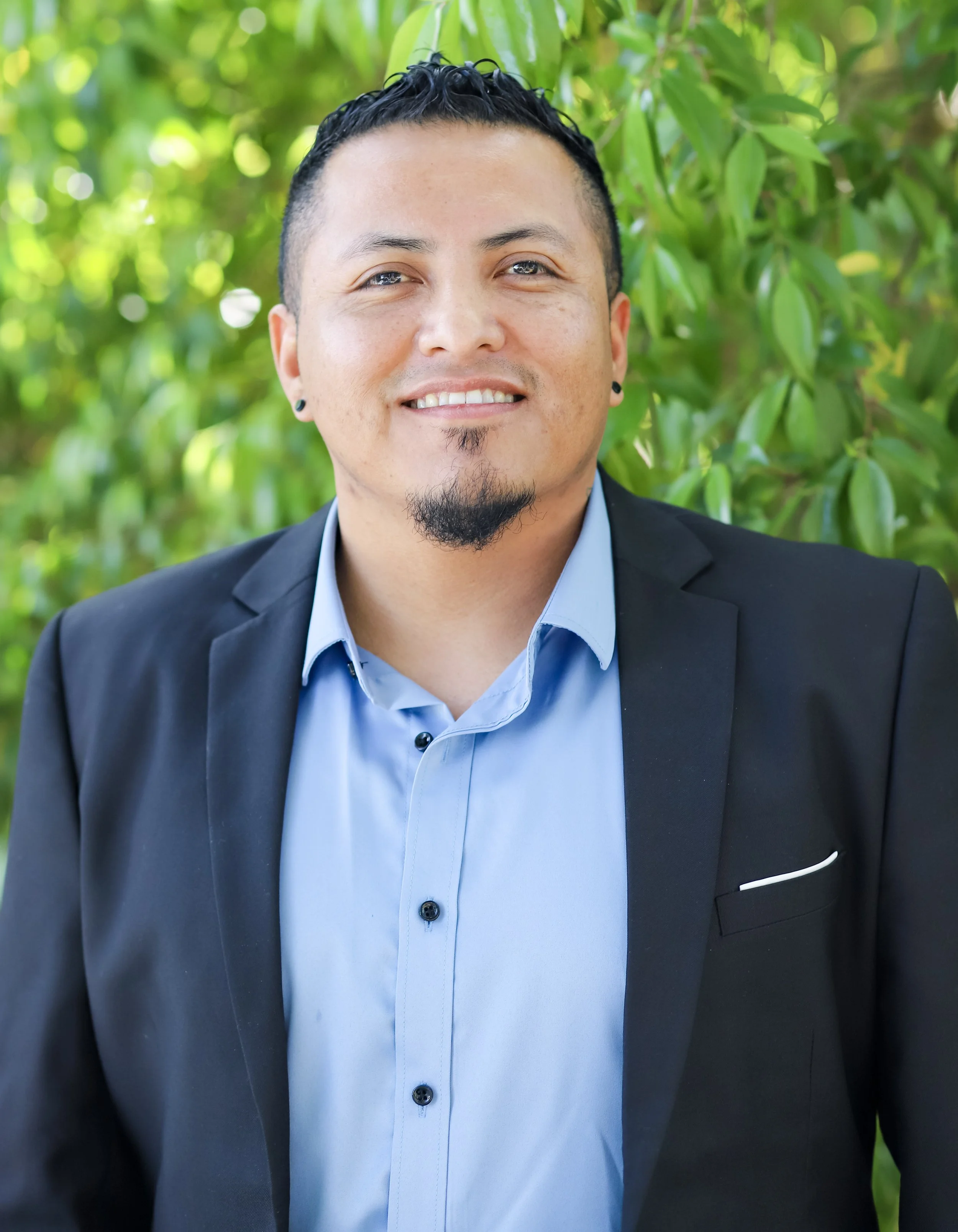 A man in a black suit jacket and blue dress shirt standing outdoors with green foliage in the background.