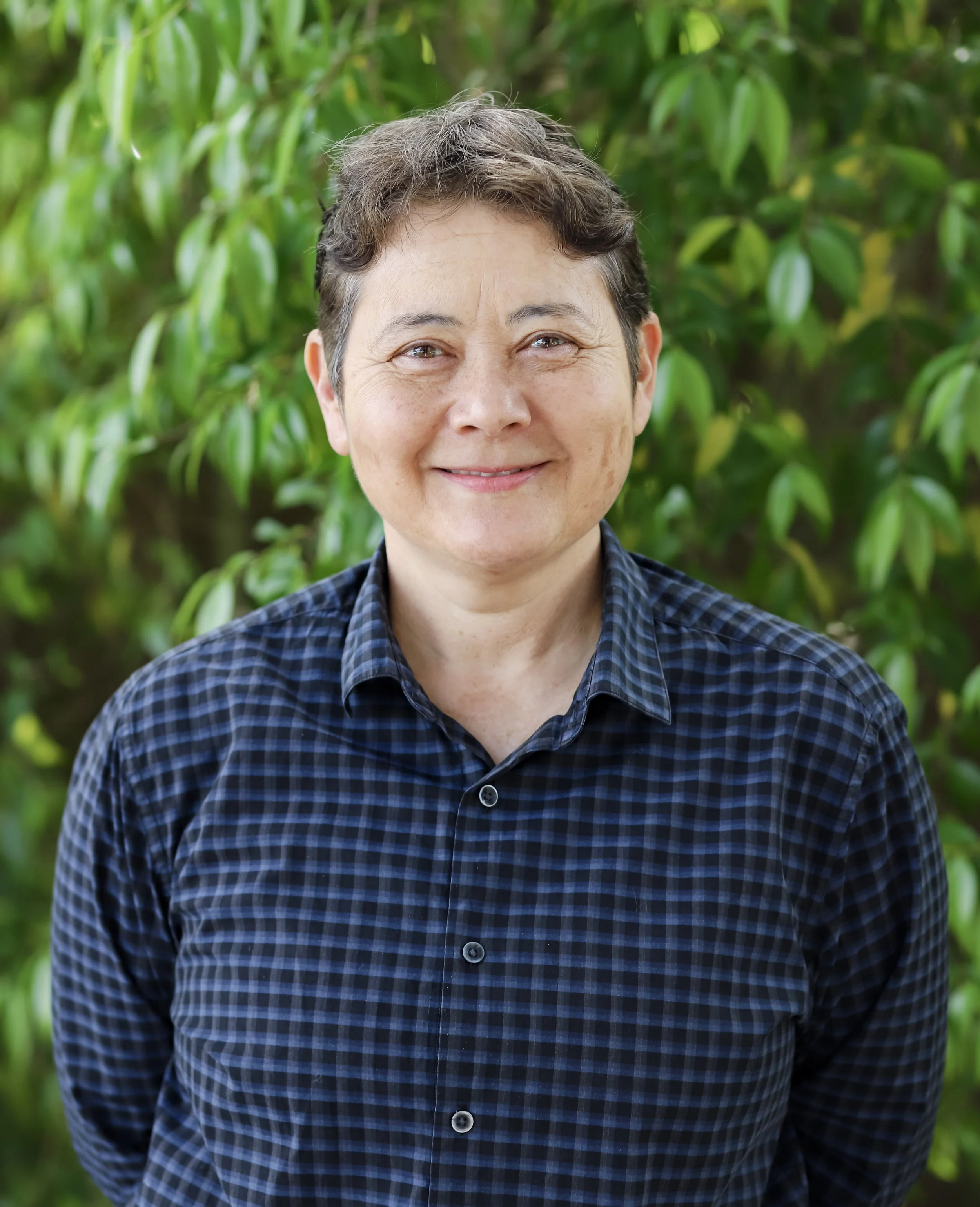 A middle-aged woman with short brown hair wearing a blue plaid shirt, standing outdoors in front of green leafy bushes, smiling at the camera.
