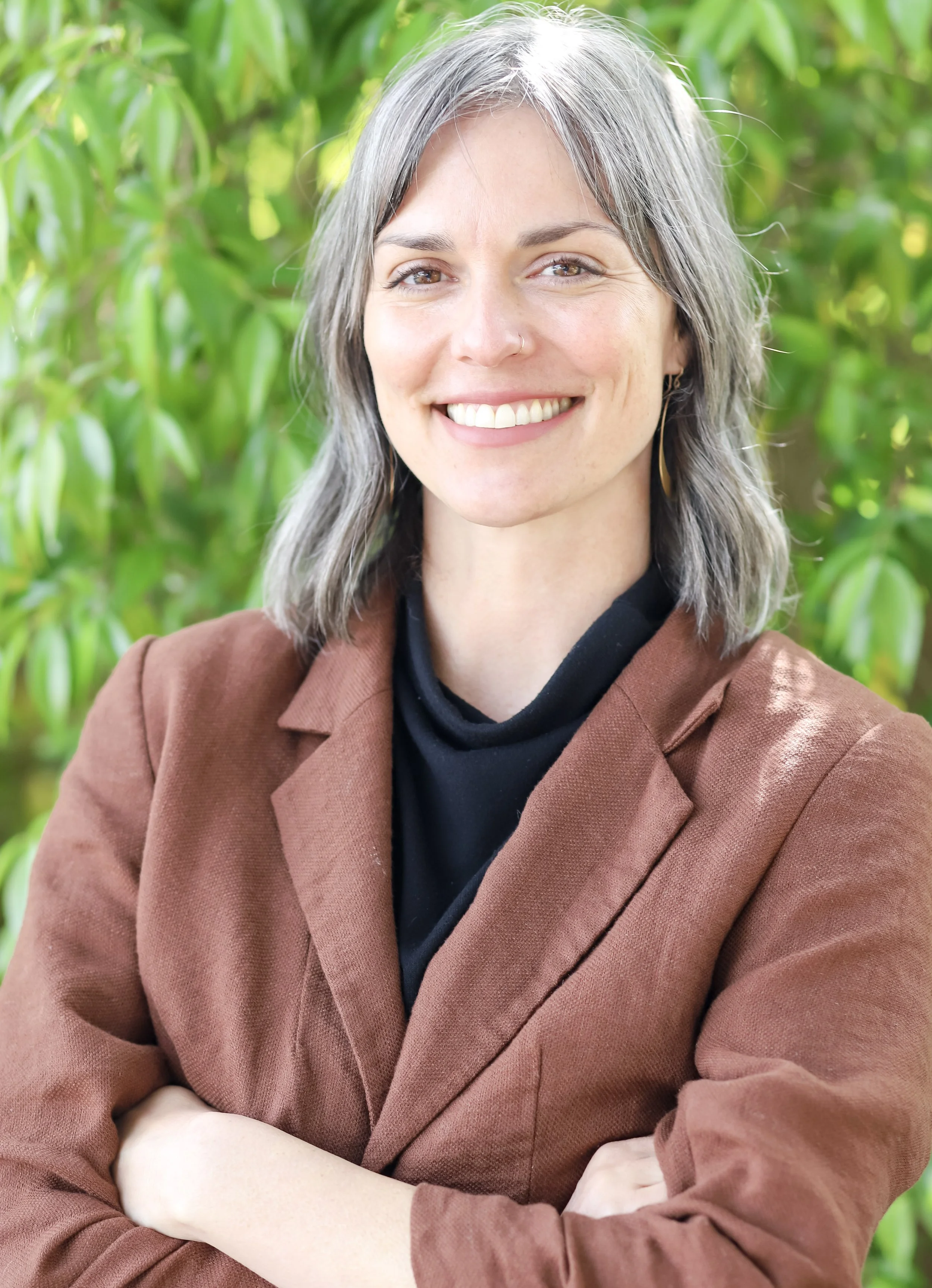 A woman with shoulder-length gray hair, smiling, standing outdoors in front of green foliage. She is wearing a brown blazer and a black top.