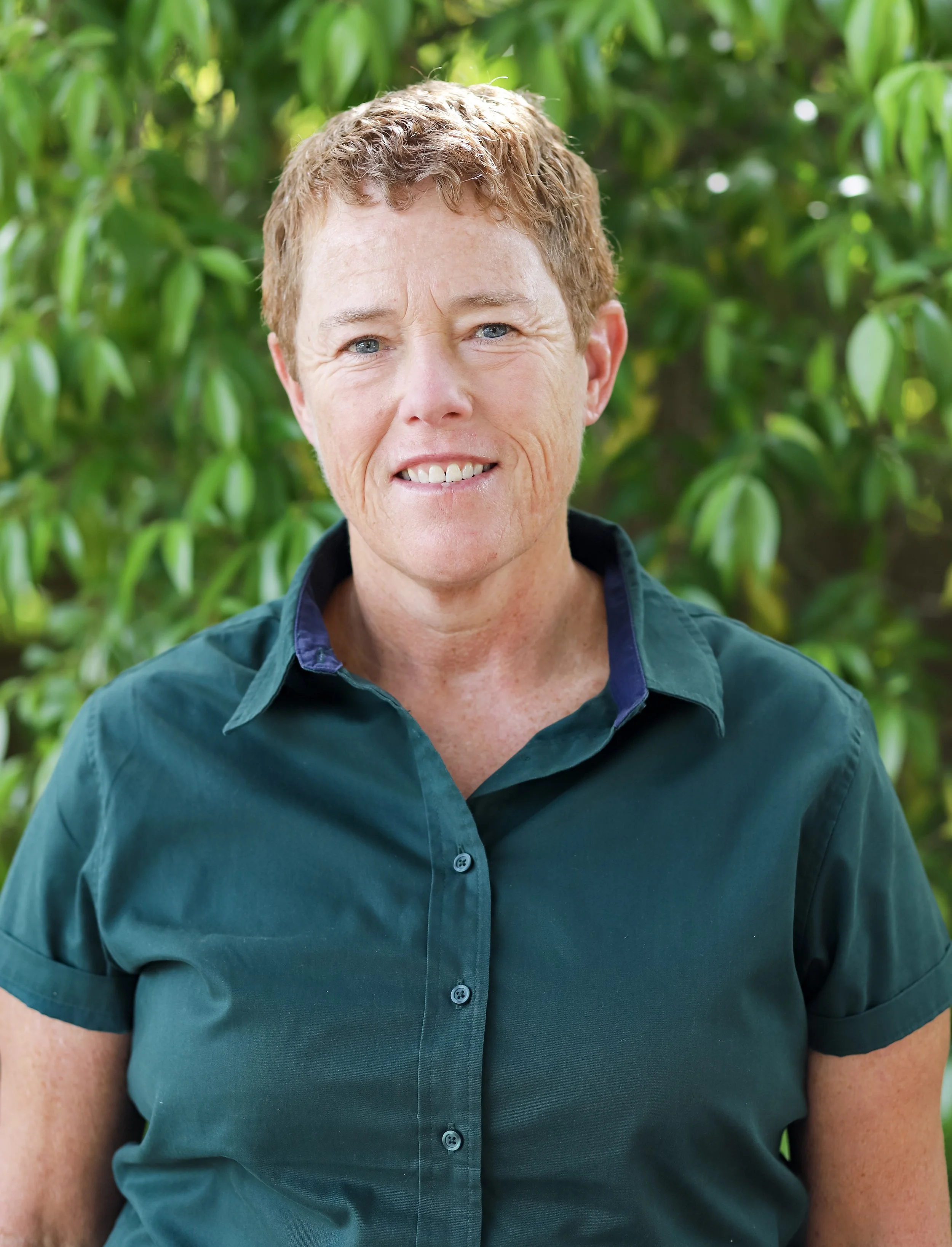 A woman with short curly red hair wearing a green button-up shirt standing outdoors in front of green foliage.