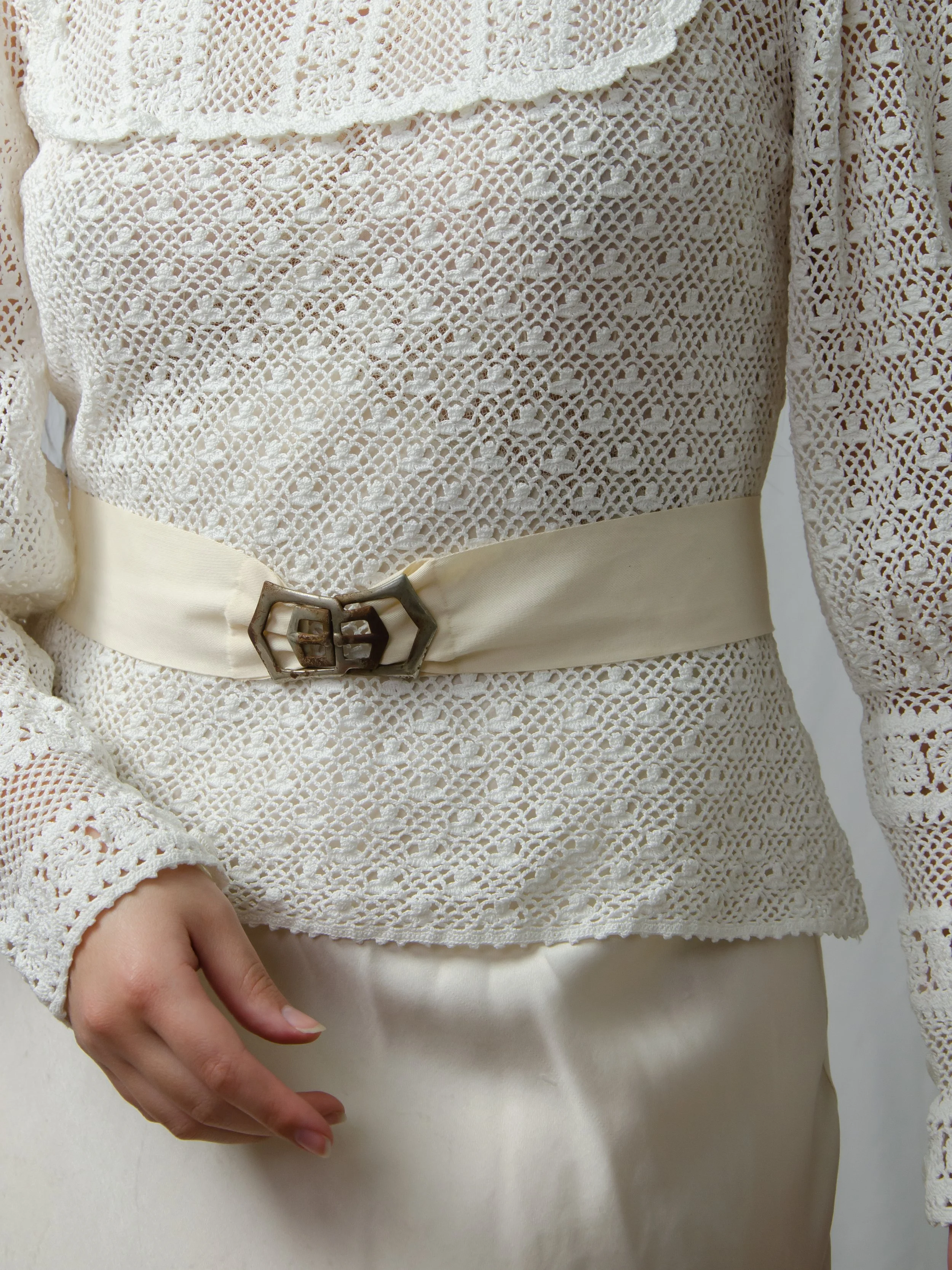 1940s ecru cotton ribbon belt with metal interlocking buckle, worn at the waist over a white crochet top.
