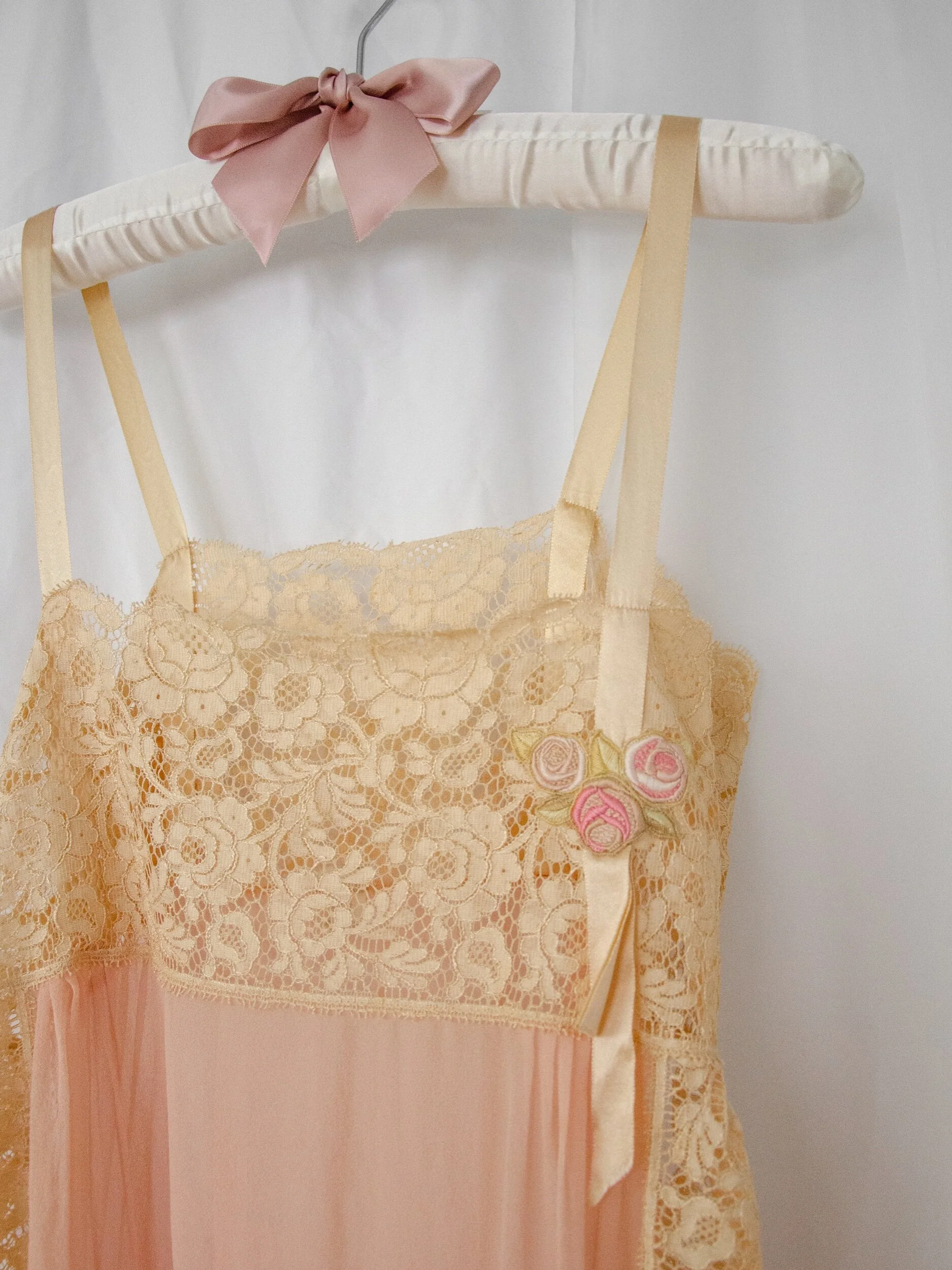 Close-up of the 1920s silk and lace teddy on a padded hanger, showing ribbon straps and rosette appliqué.